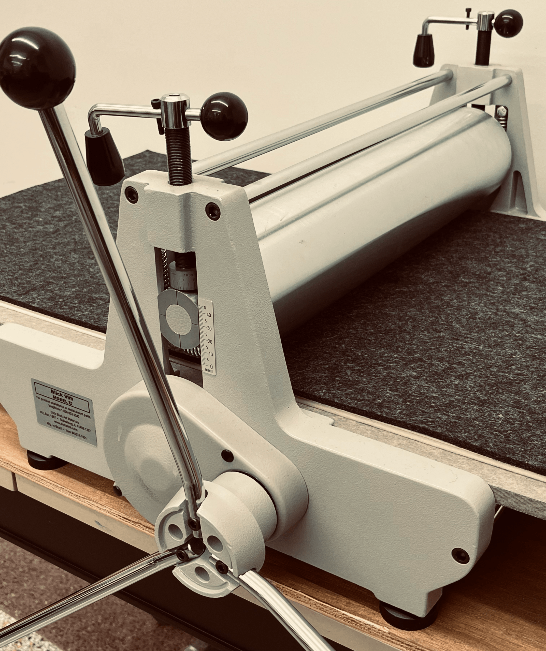 A close-up view of a printing press with large metal rollers, black handles, and adjustment levers, sitting on a felt-covered surface in a workshop setting.