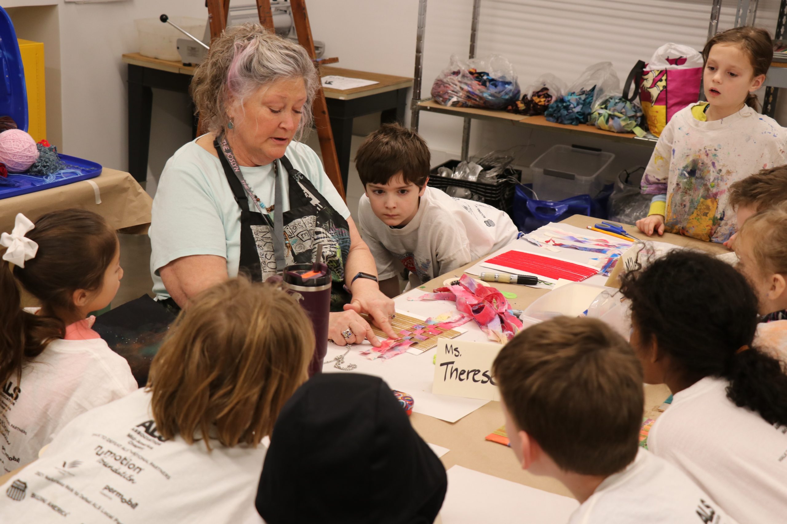 An older woman teaches a group of young children seated around a table covered with art supplies. The woman is talking as the children listen attentively. A sign on the table reads “Ms. Theresa.”.