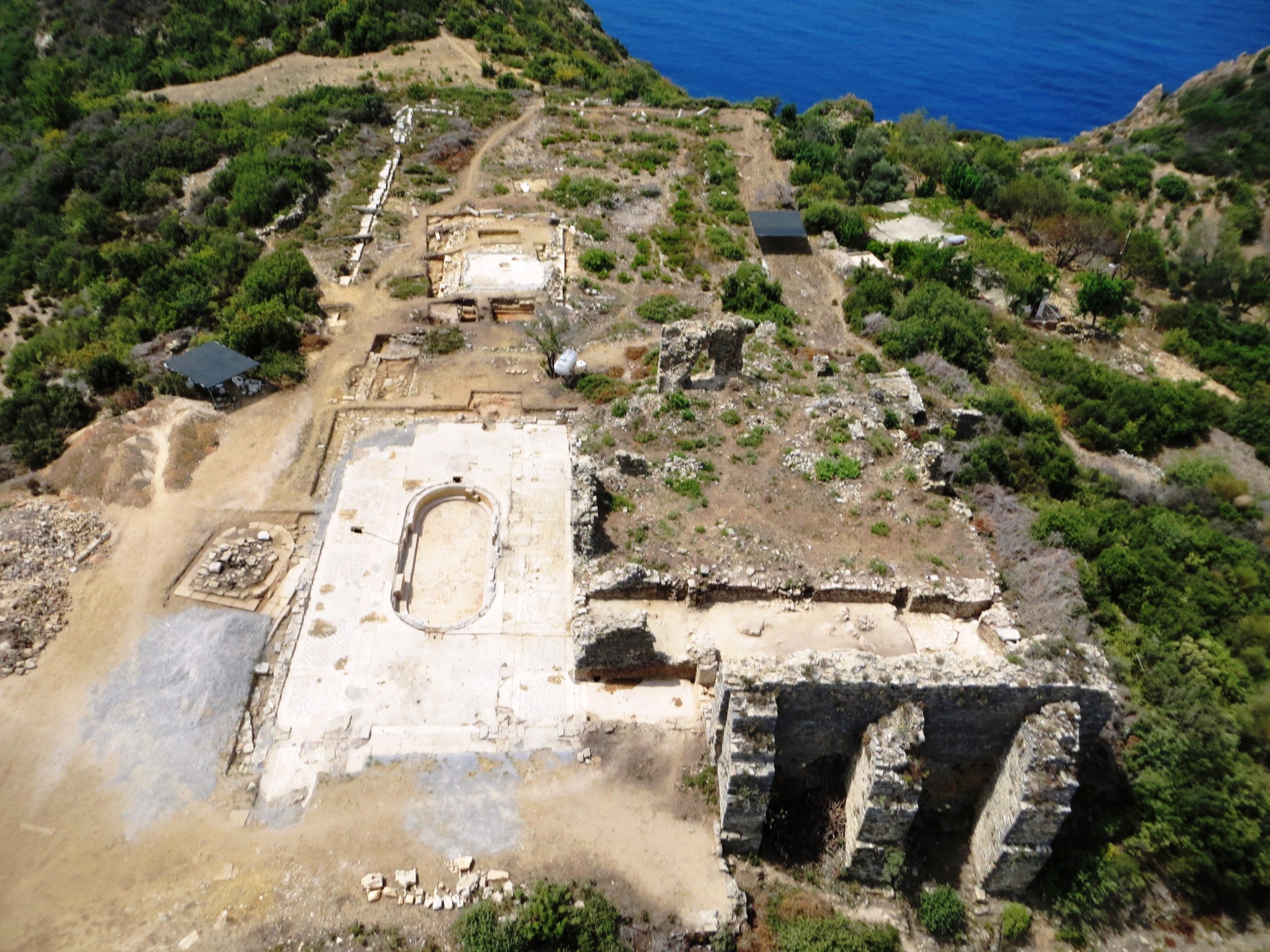 Aerial view of ancient stone ruins surrounded by greenery on a hillside, overlooking the blue sea. The site includes rectangular foundations, a central oval structure, and partially standing walls.