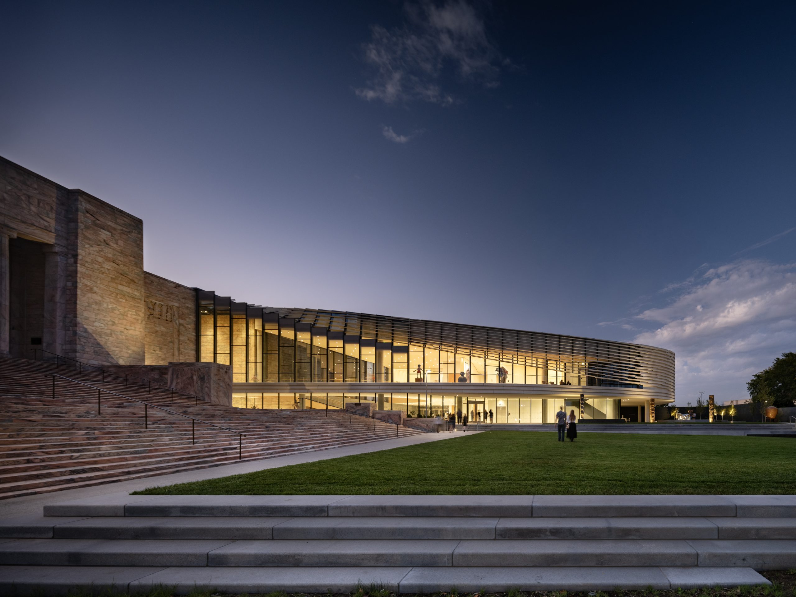 A modern building with large glass windows and curved architecture is illuminated at dusk, with a grassy lawn and steps in the foreground and two people standing near the entrance.