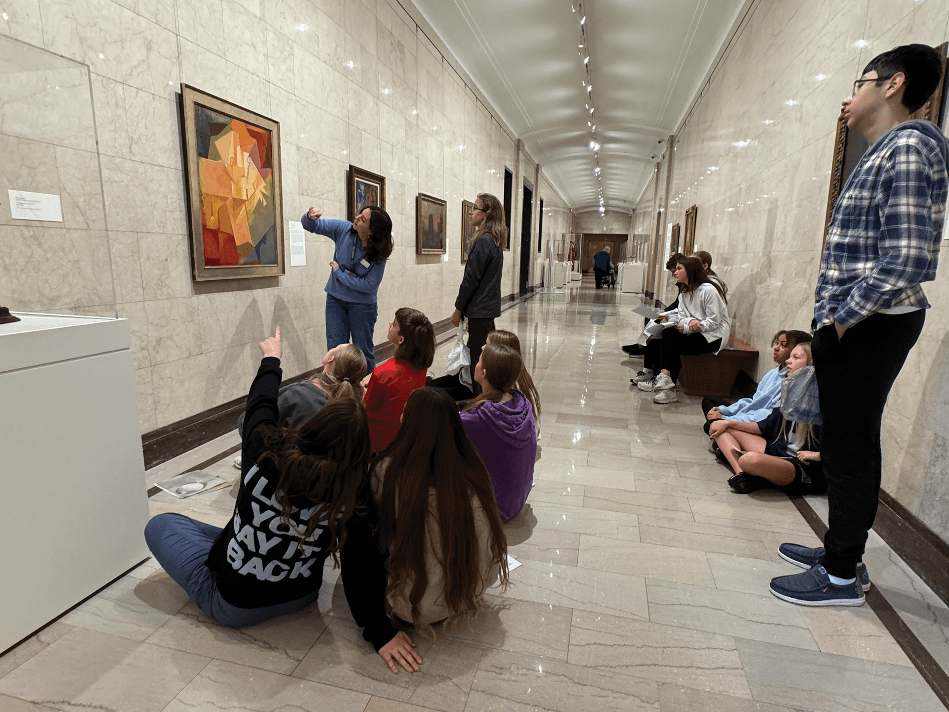 A group of students sits on the floor and benches in a museum hallway, listening to a guide who points at a colorful abstract painting on the wall. Other artworks are displayed along the marble walls.