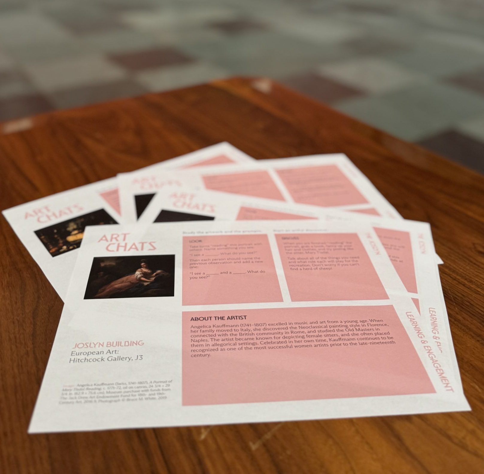 A stack of informational flyers titled "Art Chats" sits on a wooden table in an art gallery, with blurred framed paintings displayed on the pink walls in the background.