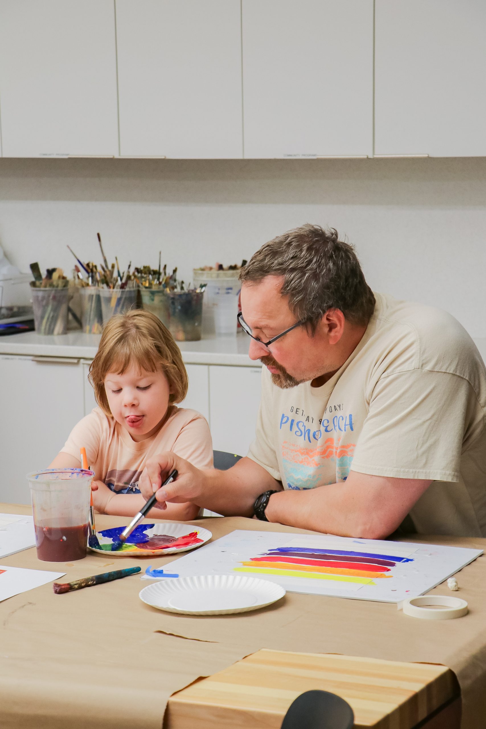 An adult and a child sit at a table painting together. The child sticks out their tongue in concentration while using a paintbrush, and the adult assists, both surrounded by art supplies.
