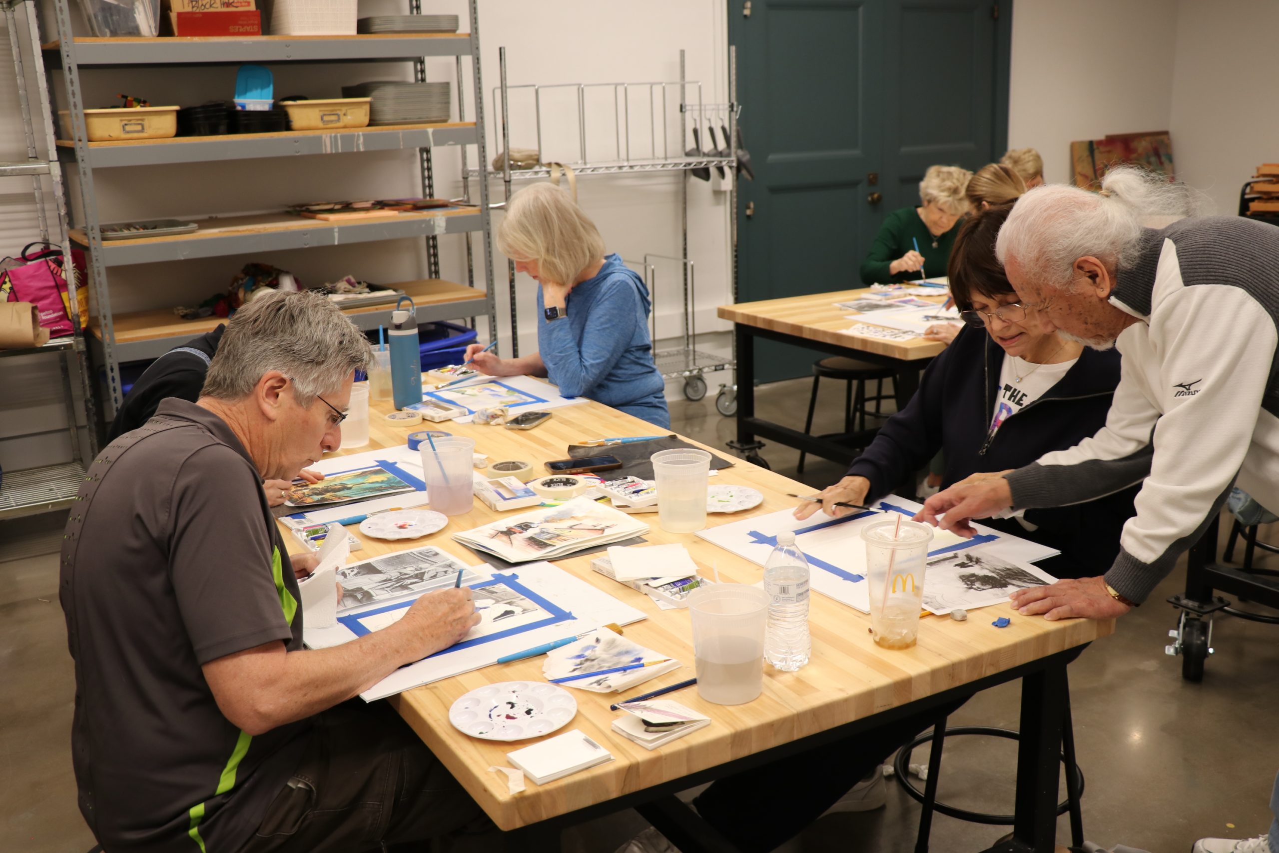 A group of older adults sit around tables in an art classroom, painting and drawing. One man stands, assisting a seated woman, while others focus on their own art projects. Art supplies are scattered on the tables.