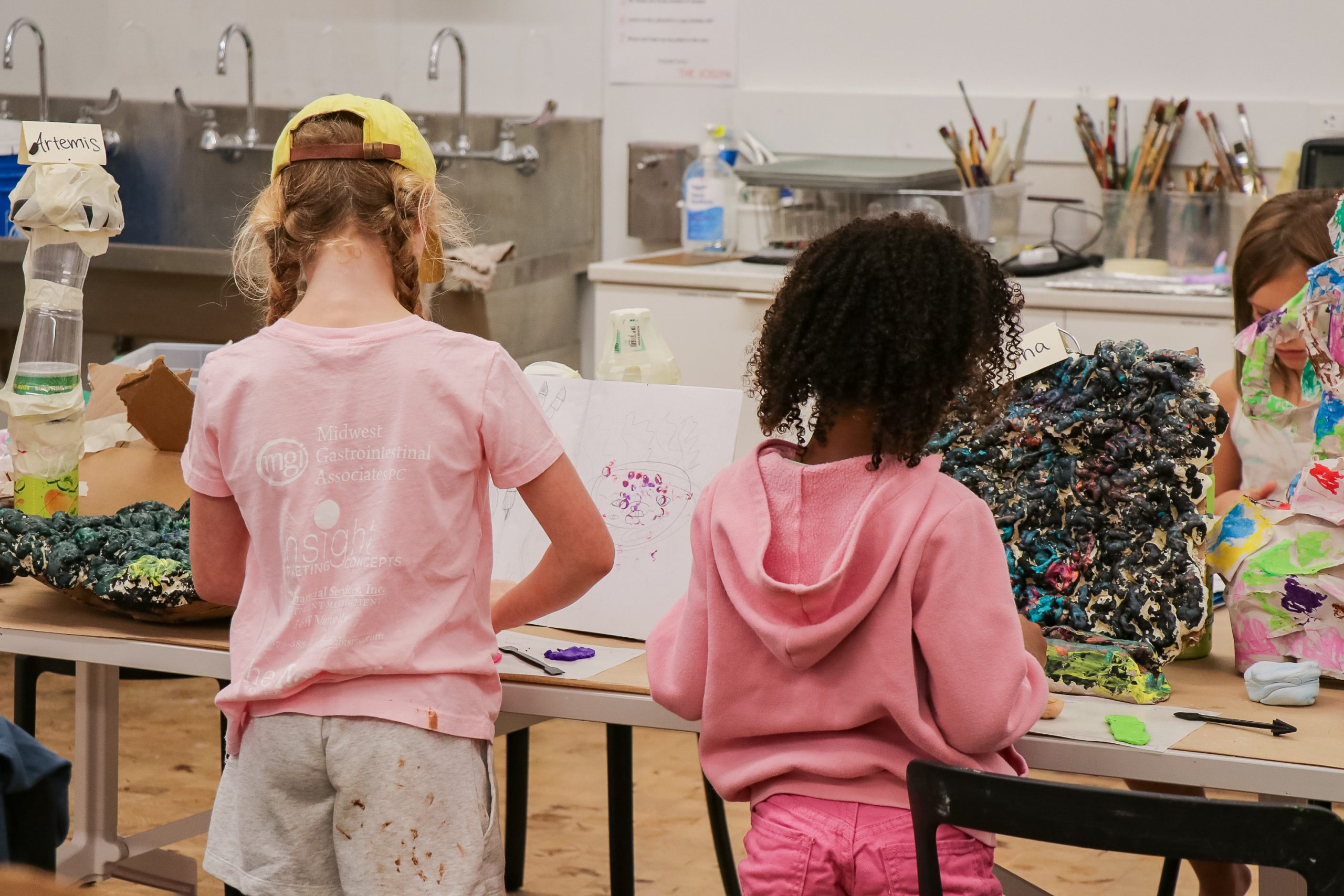Two children with curly hair, dressed in pink, stand at a table covered with colorful art supplies and sculptures in a classroom or art studio. Sinks, brushes, and cleaning supplies are visible in the background.
