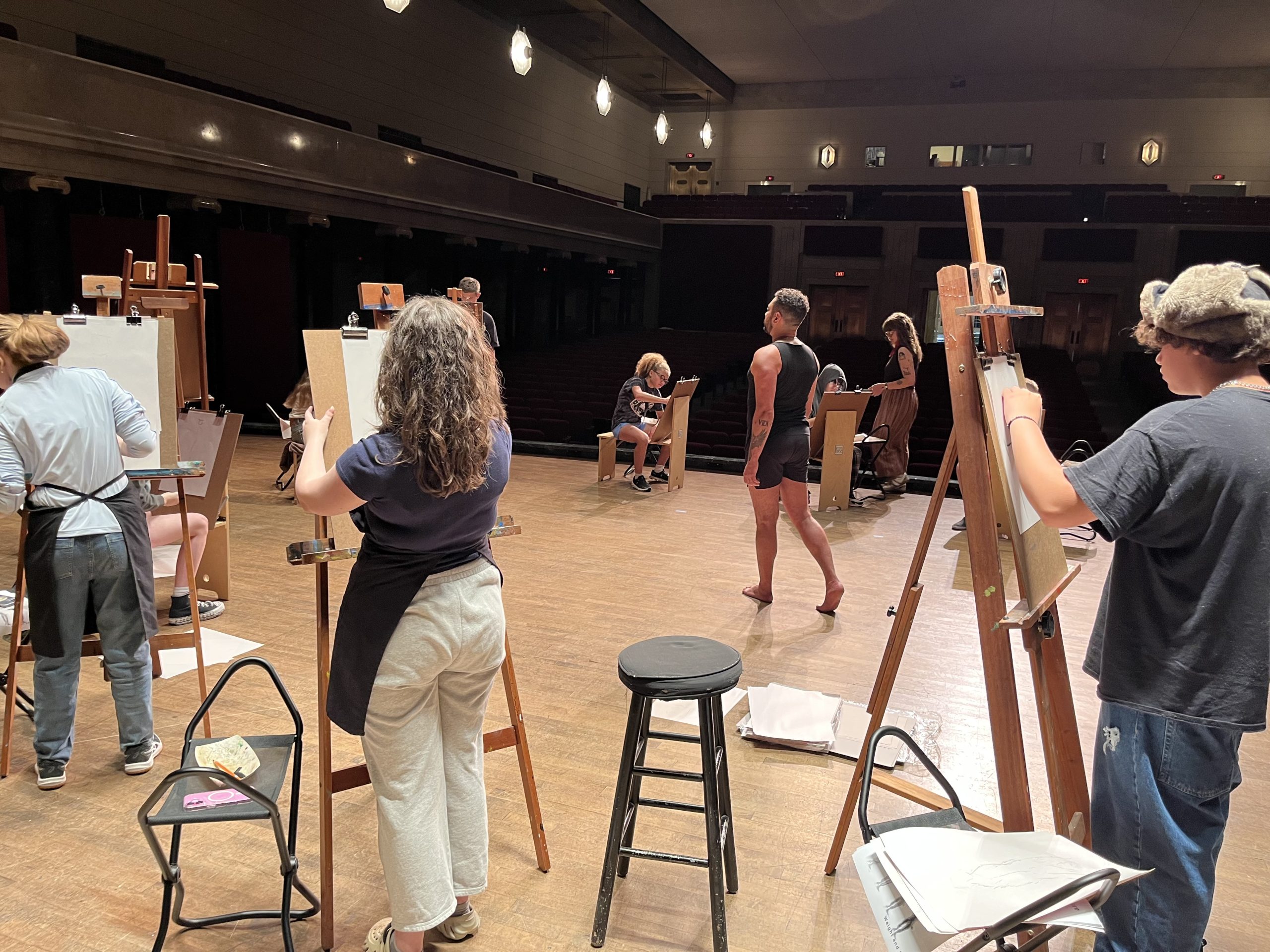 Several artists stand at easels sketching a person posing on a platform in the center of a large, well-lit room that appears to be a theater or auditorium. Art supplies and stools are scattered around.