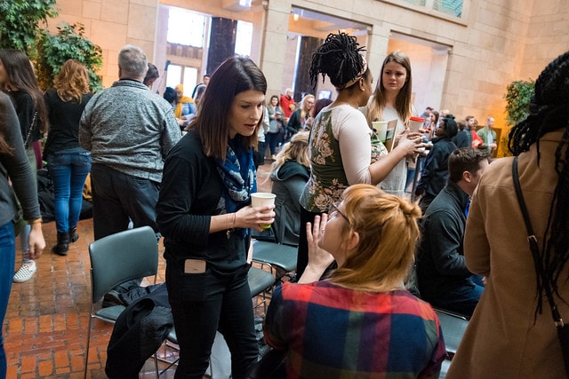 A group of people socializes indoors, some standing and others sitting, many holding cups. The setting has brick flooring and high ceilings, with natural light streaming in and plants along the walls.