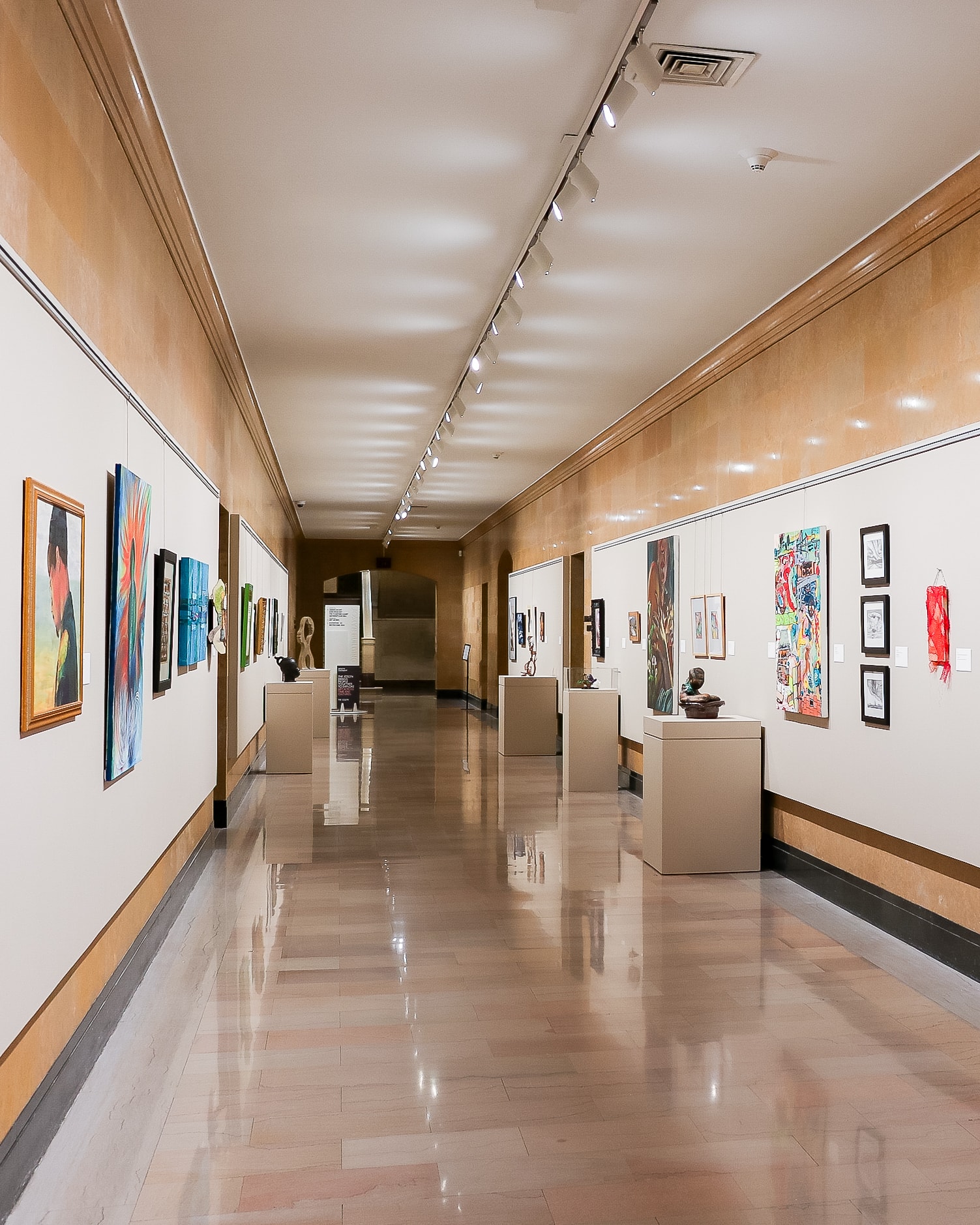 A well-lit art gallery hallway with polished floors, displaying colorful paintings and sculptures on walls and pedestals, creating a modern and inviting atmosphere.