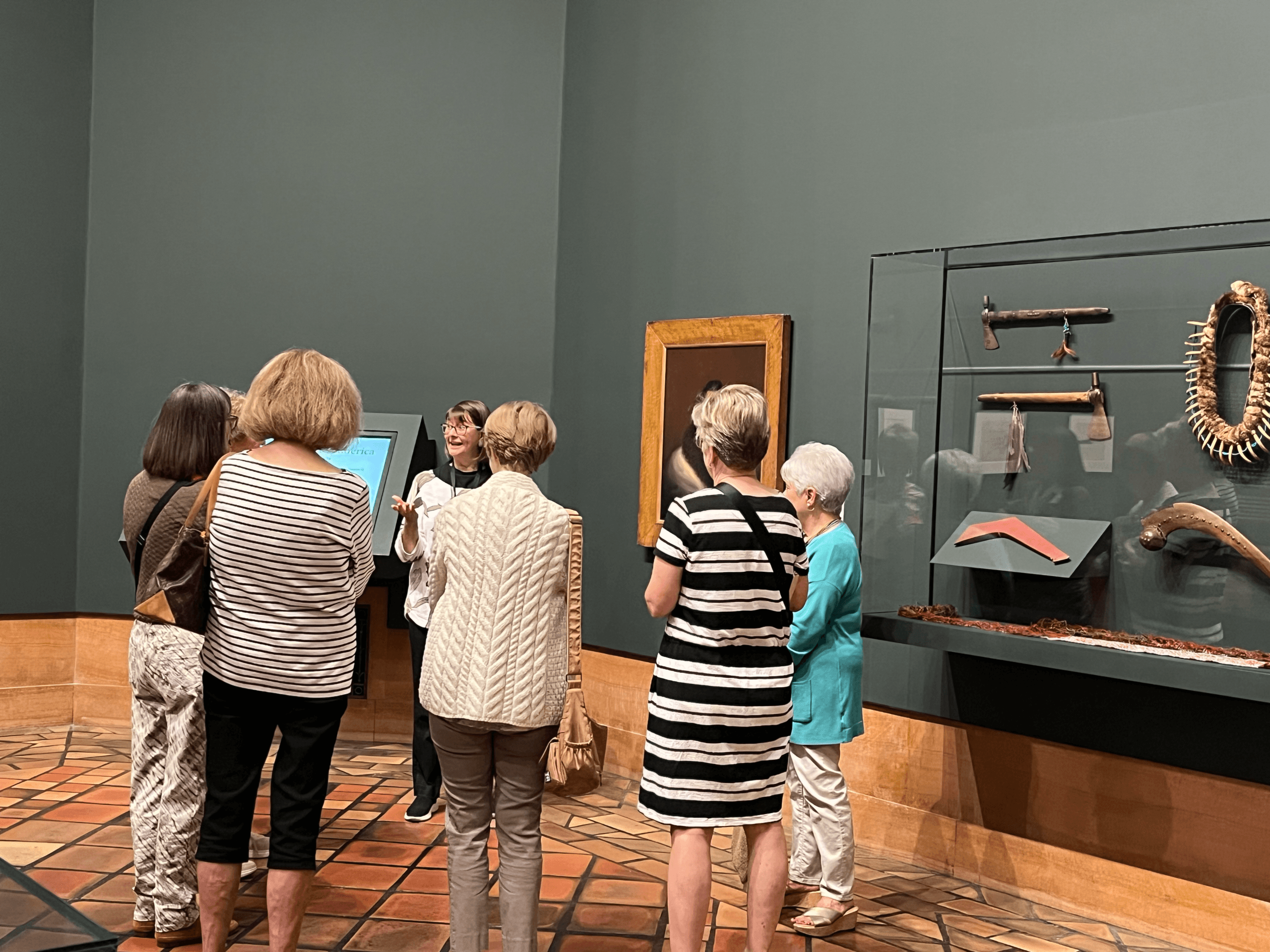 A group of women stand in an art museum, listening to a guide. They face display cases with cultural artifacts and tools mounted on the wall. The room has dark green walls and patterned tile flooring.