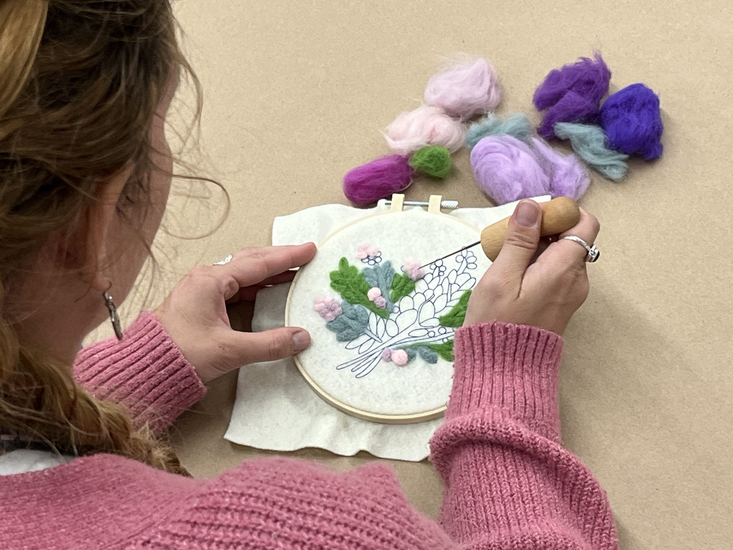 A person wearing a pink sweater uses a needle to embroider colorful wool onto fabric in a hoop, with bundles of pink, purple, blue, and green wool nearby on a beige surface.