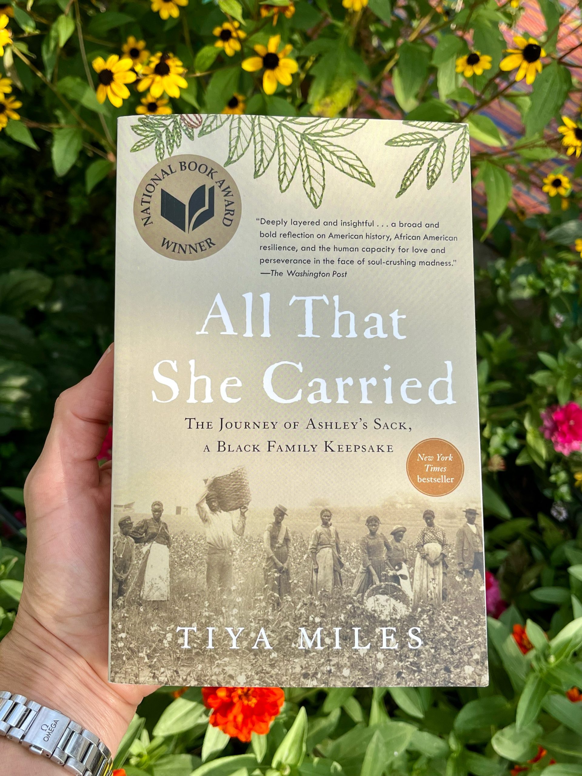 A hand holds the book "All That She Carried" by Tiya Miles, featuring a sepia photo of Black women and children on the cover, against a backdrop of blooming flowers and green leaves outdoors.