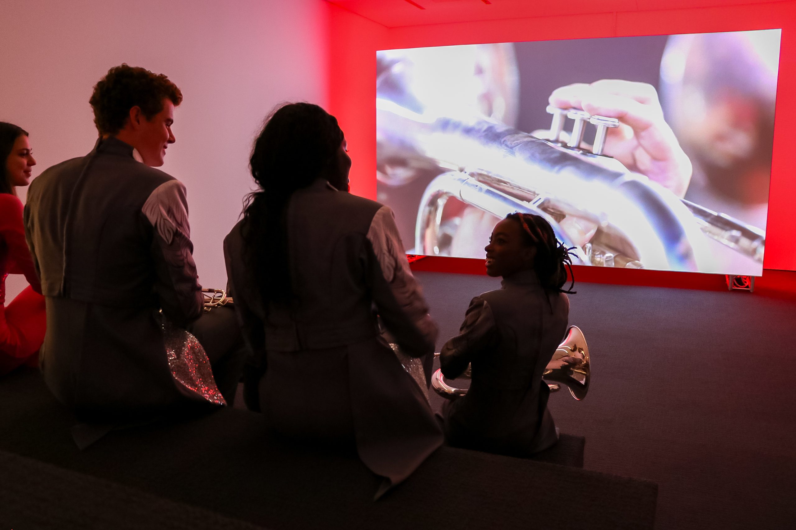 Four people in matching gray outfits sit in a dark room, facing a large screen showing a close-up of someone playing a brass instrument. The scene is illuminated by red light from the screen.