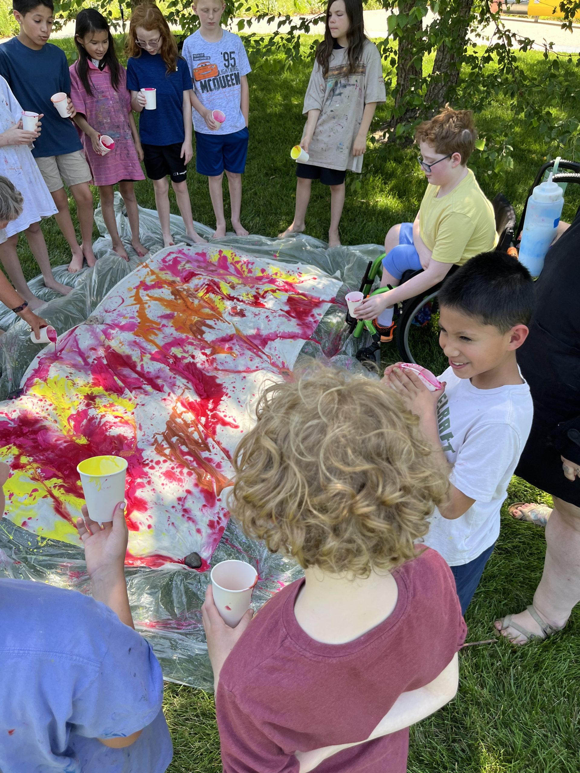 A group of children stand around a large plastic sheet on grass, covered in colorful splatters from cups they hold, enjoying an outdoor art activity in the shade.