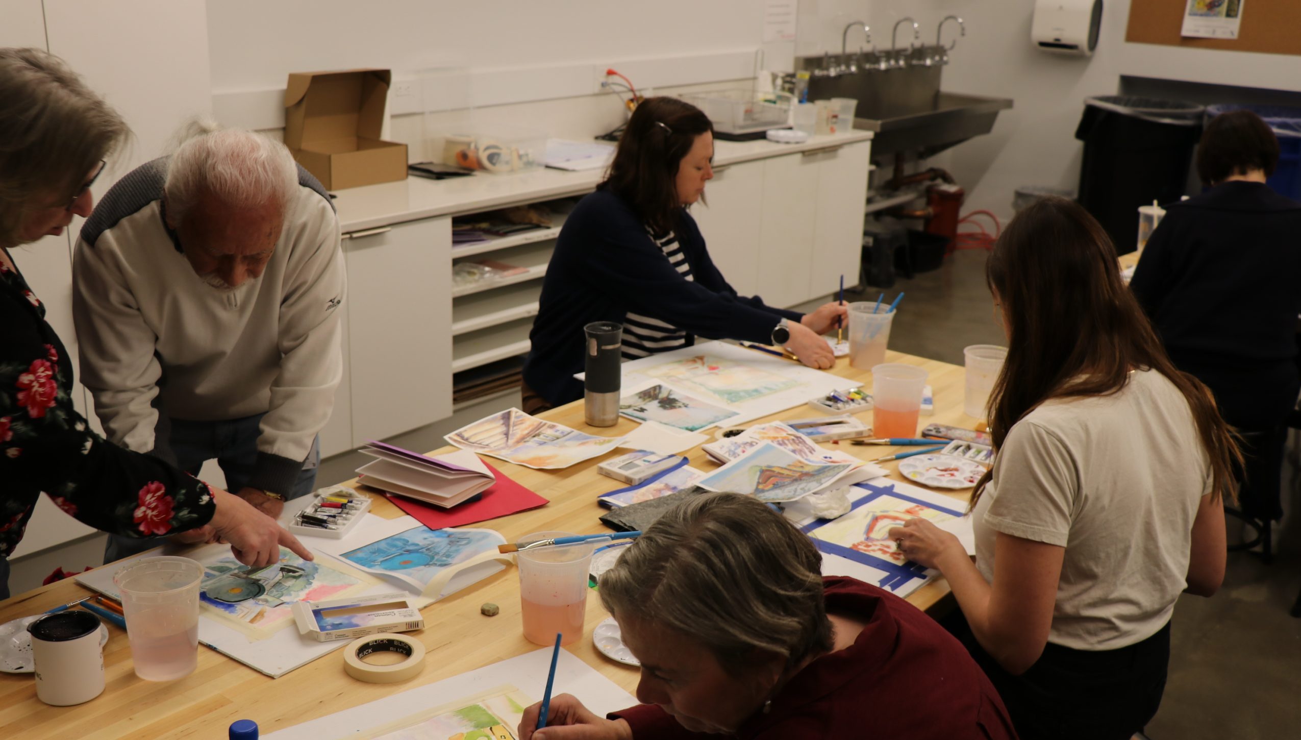 Five adults sit and stand around a table covered with art supplies, paper, and drinks, as they focus on painting and drawing in a bright, modern art classroom.