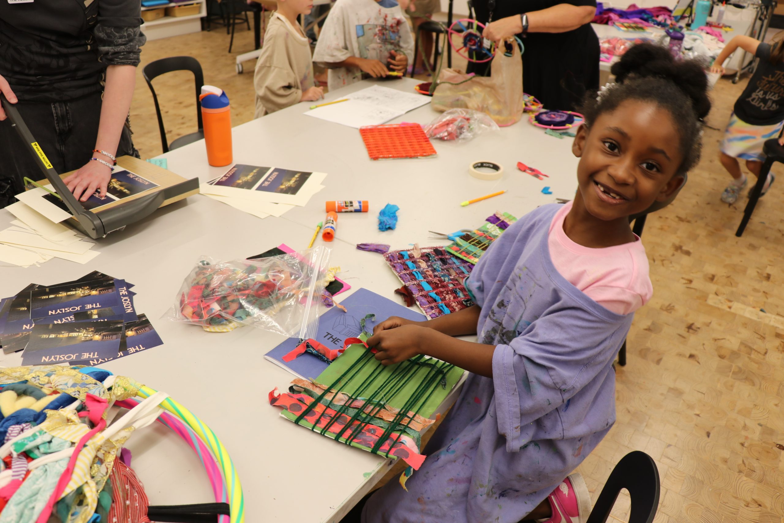 A smiling young girl weaves colorful fabric strips on a loom at a table covered with art supplies, surrounded by other children and adults in a busy, creative indoor setting.