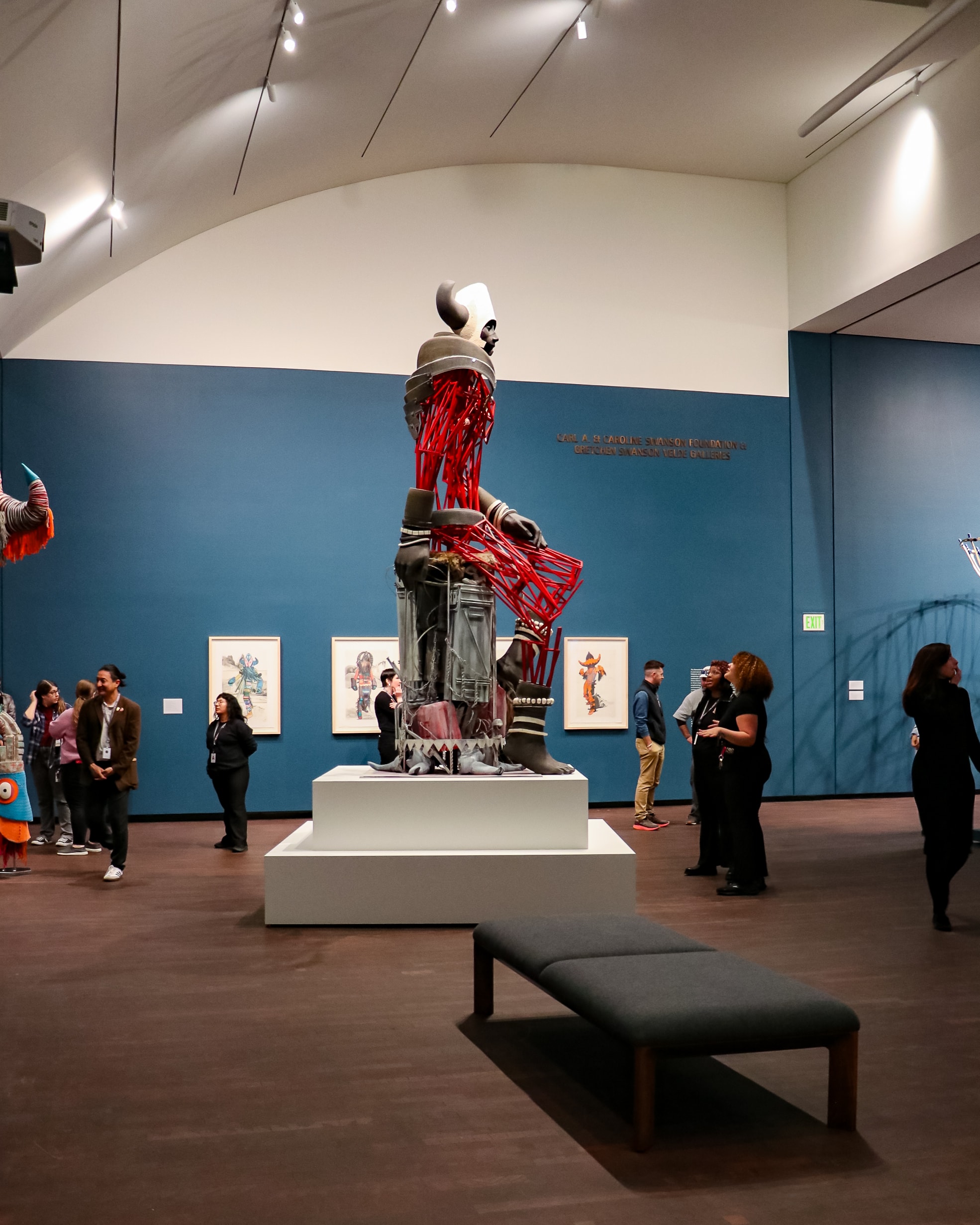People observe a large, colorful sculpture with red, gray, and white elements in an art gallery. Several framed artworks hang on the blue walls, and a gray bench is placed in the foreground.