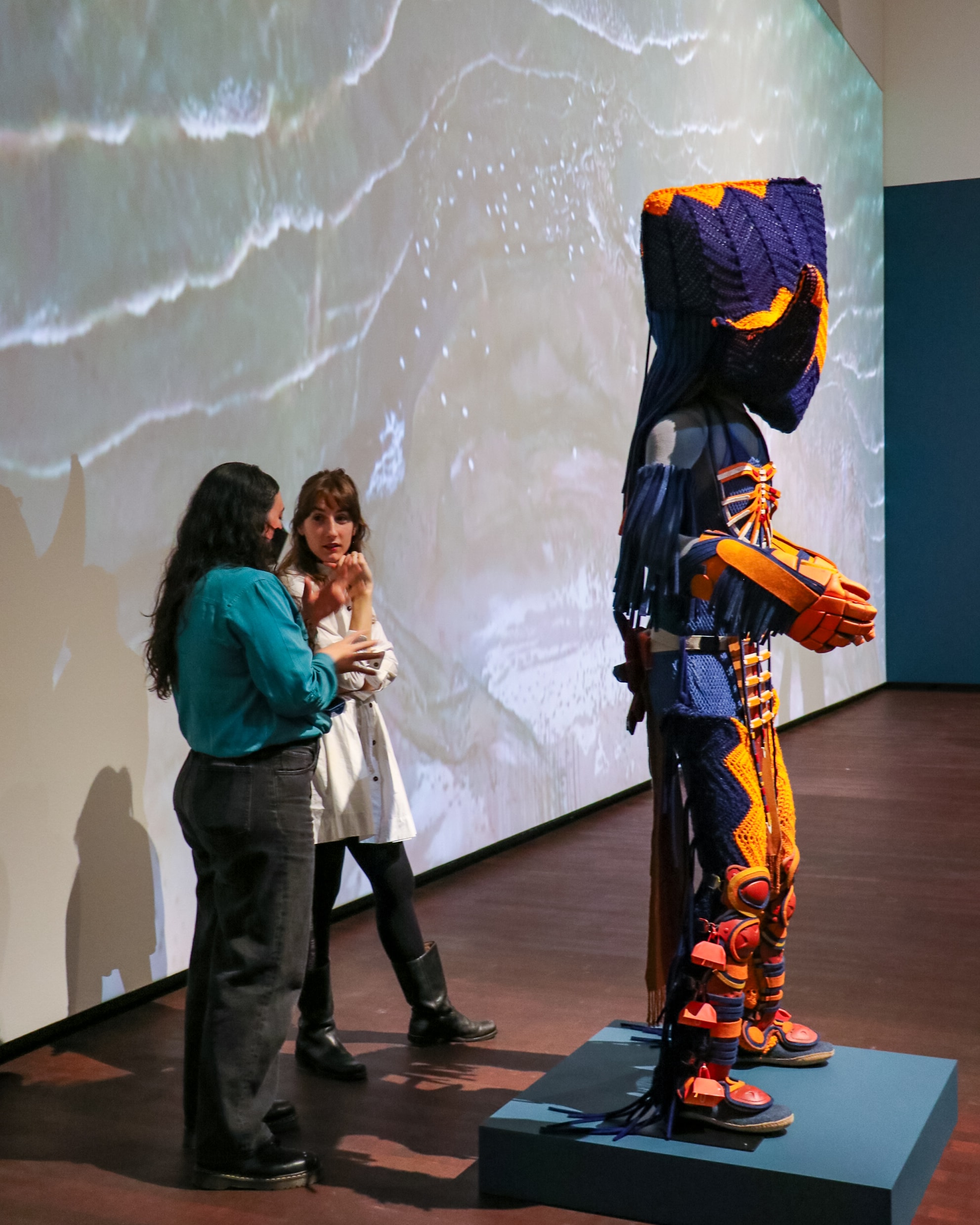 Two people stand in a gallery, talking in front of a large, colorful, abstract humanoid sculpture made of woven materials. A projection of ocean waves covers the wall behind them.