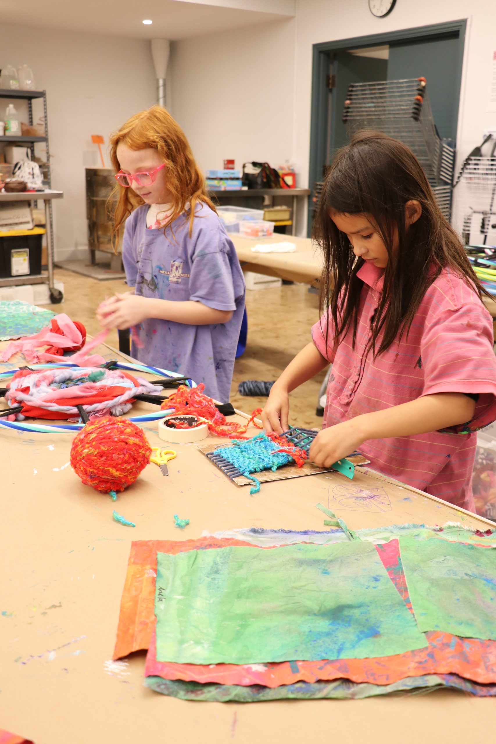 Two children work on colorful art projects at a table covered with paper, yarn, and craft materials in a classroom or art studio setting. One child wears pink, the other wears purple.