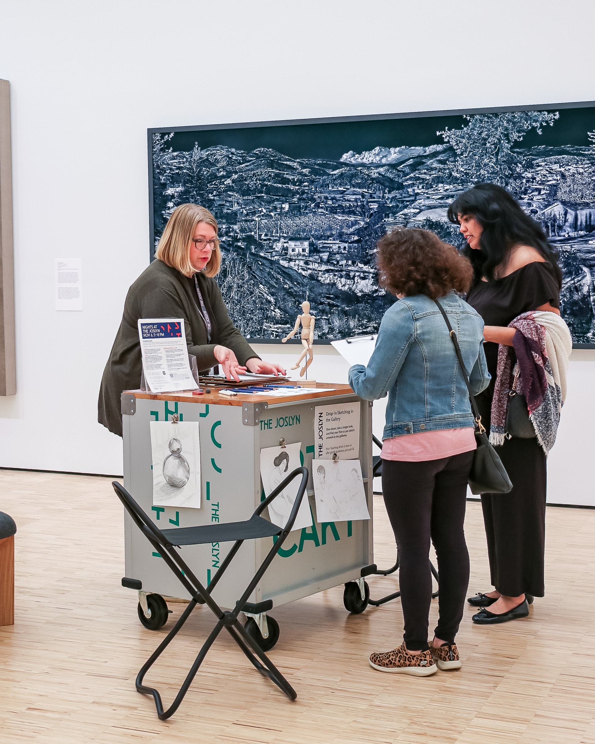 Three women stand around an art activity cart in a museum gallery, interacting with papers and materials on the cart. A detailed black-and-white artwork hangs on the wall behind them.