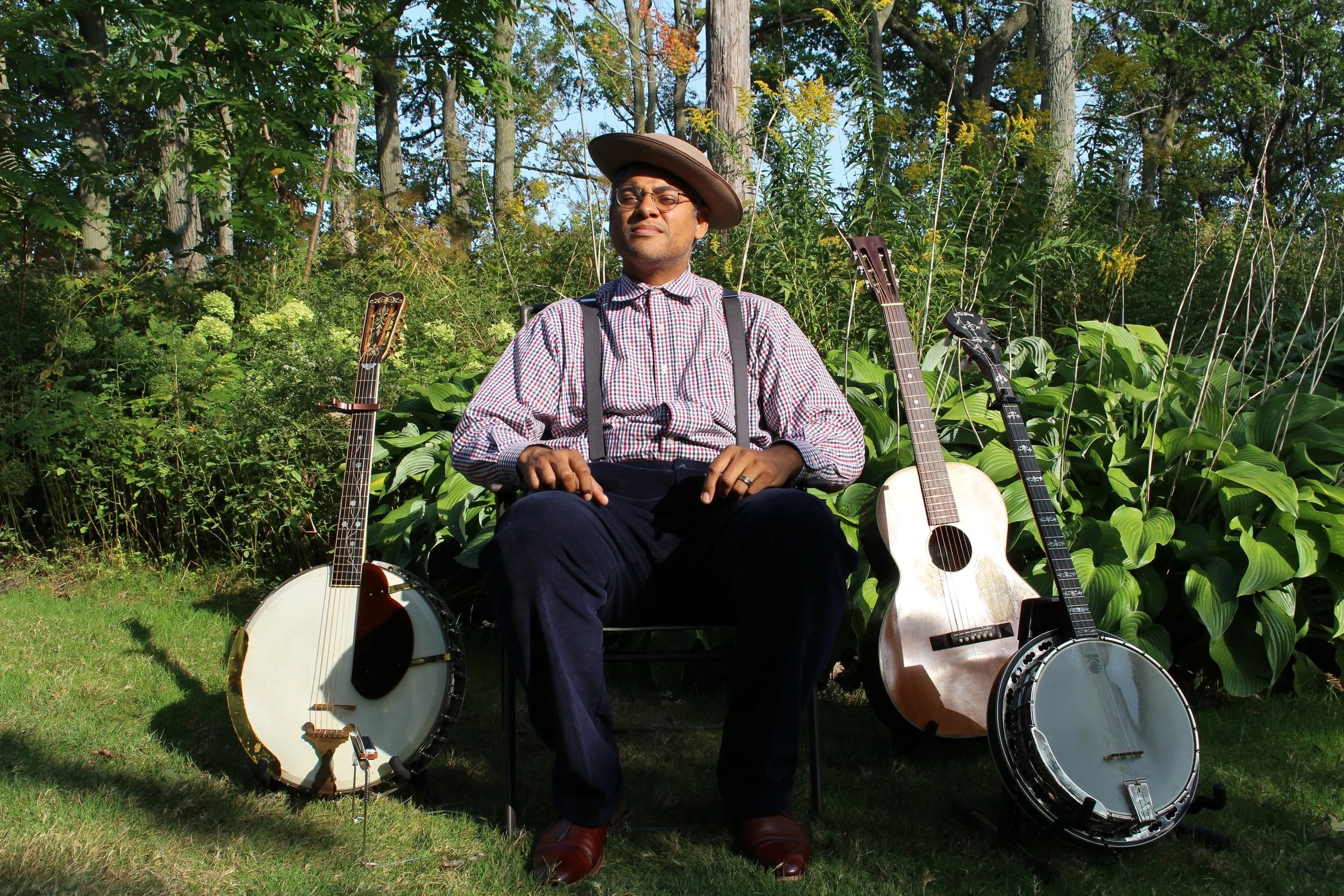 A man wearing suspenders, a checked shirt, and a hat sits outdoors on a chair between two banjos and a guitar, surrounded by green plants and trees in bright sunlight.