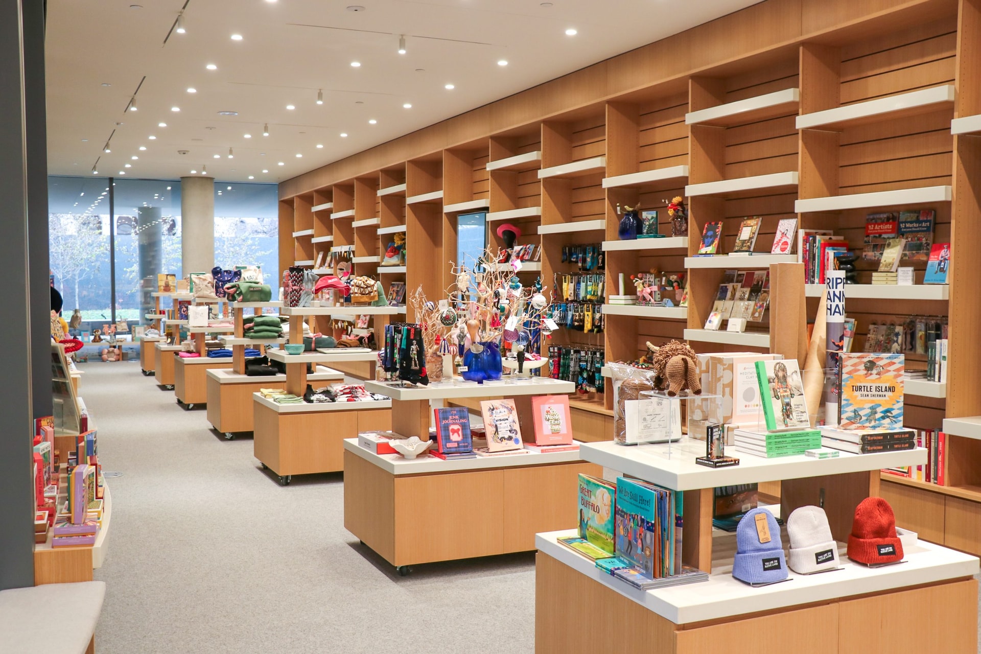 A modern bookstore with wooden shelves and display tables filled with books, toys, gifts, and souvenirs. The space is well-lit and organized, creating a welcoming shopping environment.