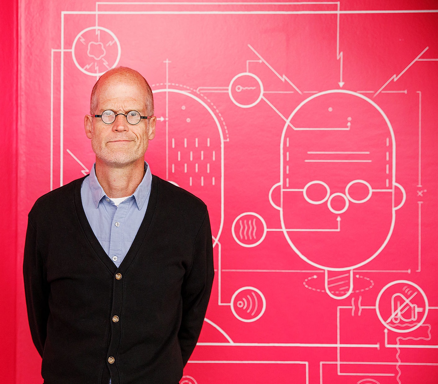 A man with glasses and a black cardigan stands in front of a bright pink wall featuring a white, abstract drawing of a face and geometric shapes.