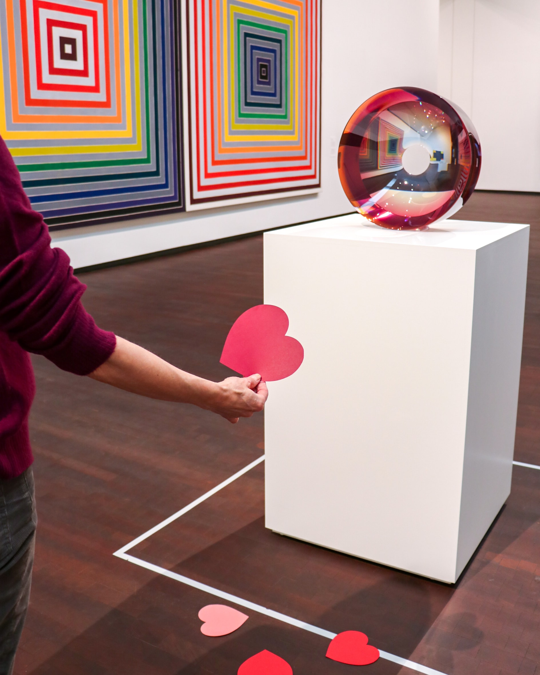 A person holding a red paper heart stands near a white pedestal displaying a round, reflective sculpture in a gallery with colorful, geometric wall art. Red paper hearts are scattered on the floor.
