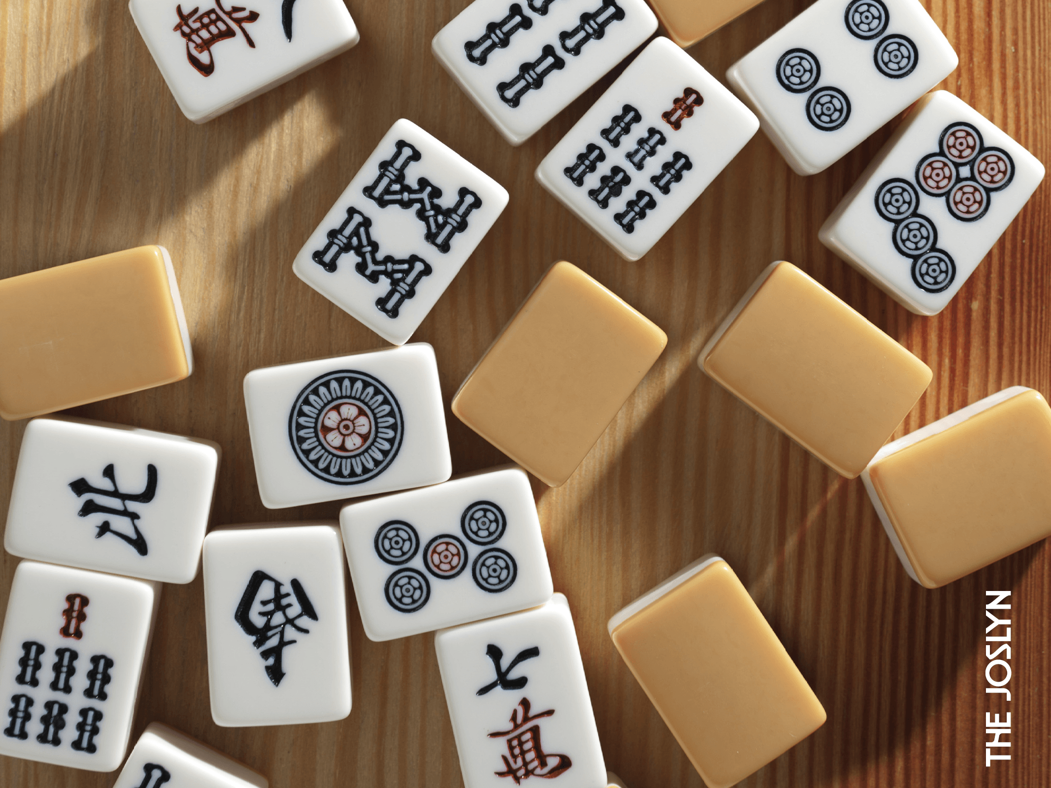 Mahjong tiles with various symbols and patterns are scattered on a wooden surface. Some tiles show circles, bamboo, and Chinese characters. The word "THE JOSLYN" appears in white text on the bottom right.