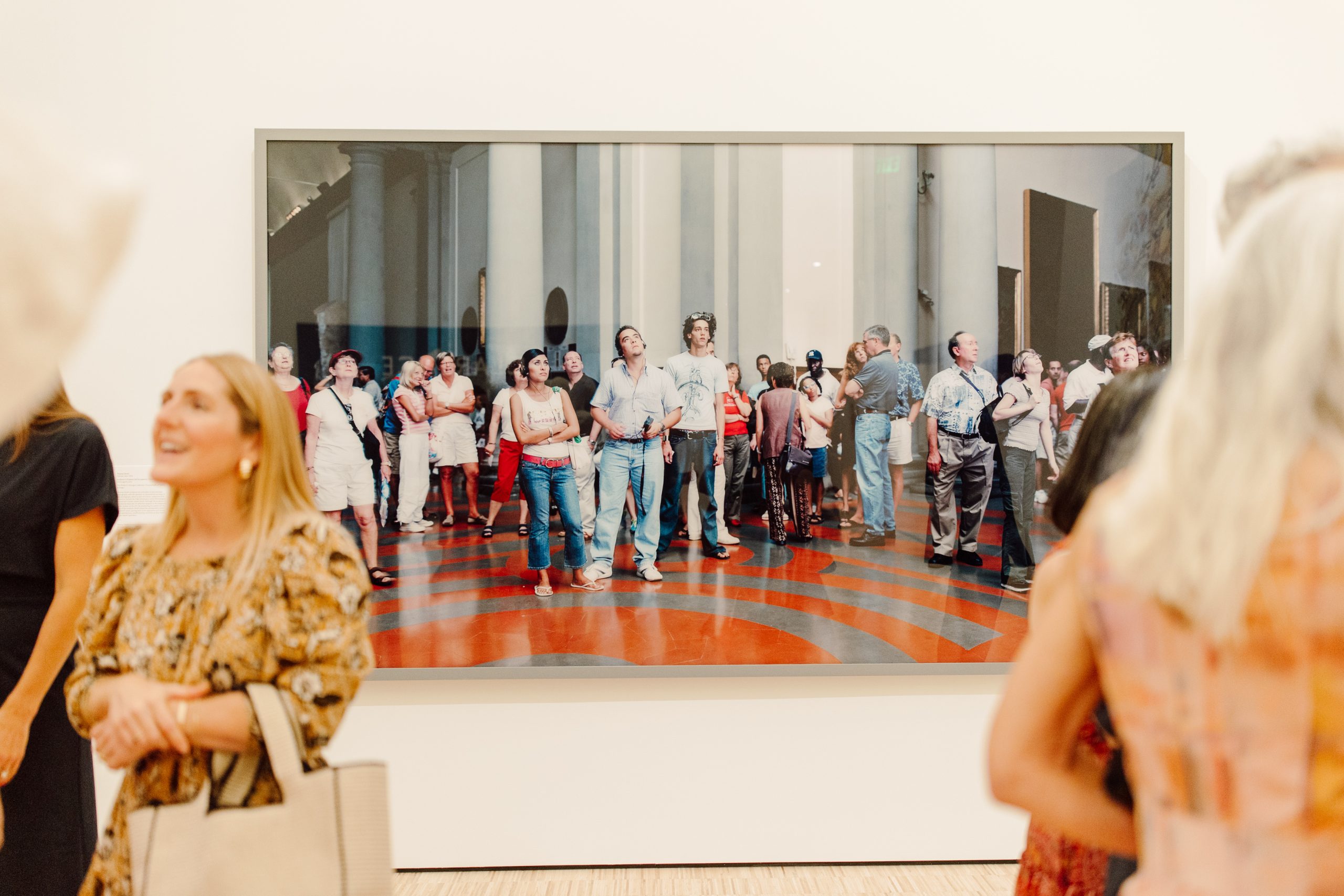 People in an art gallery stand and talk in front of a large framed photo showing a crowd inside a classical building with columns and a red and black patterned floor. The gallery visitors appear engaged and relaxed.