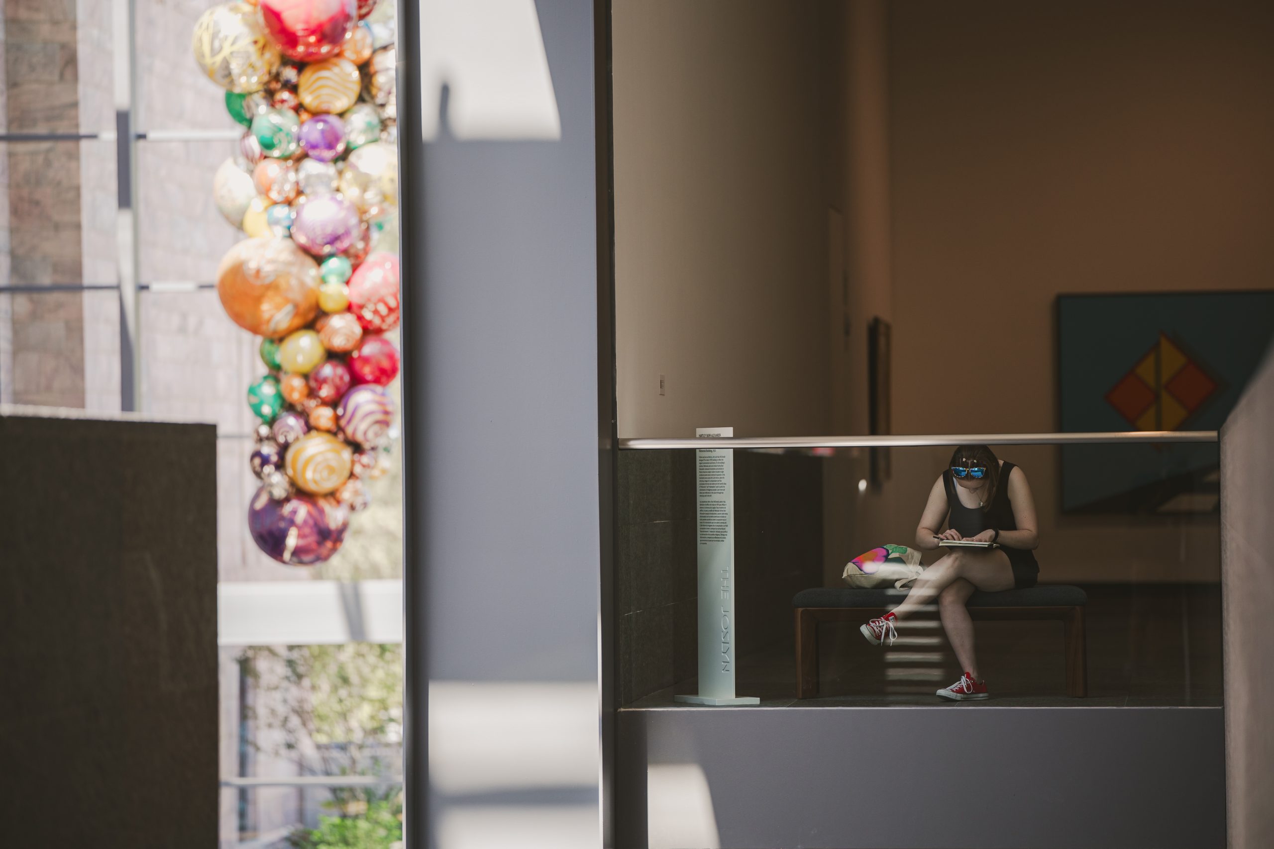 A person sits alone on a bench in a modern, sunlit building, reading. Colorful glass sculptures hang on the left, seen through a window. The scene is calm and quiet, with geometric shadows and natural light.