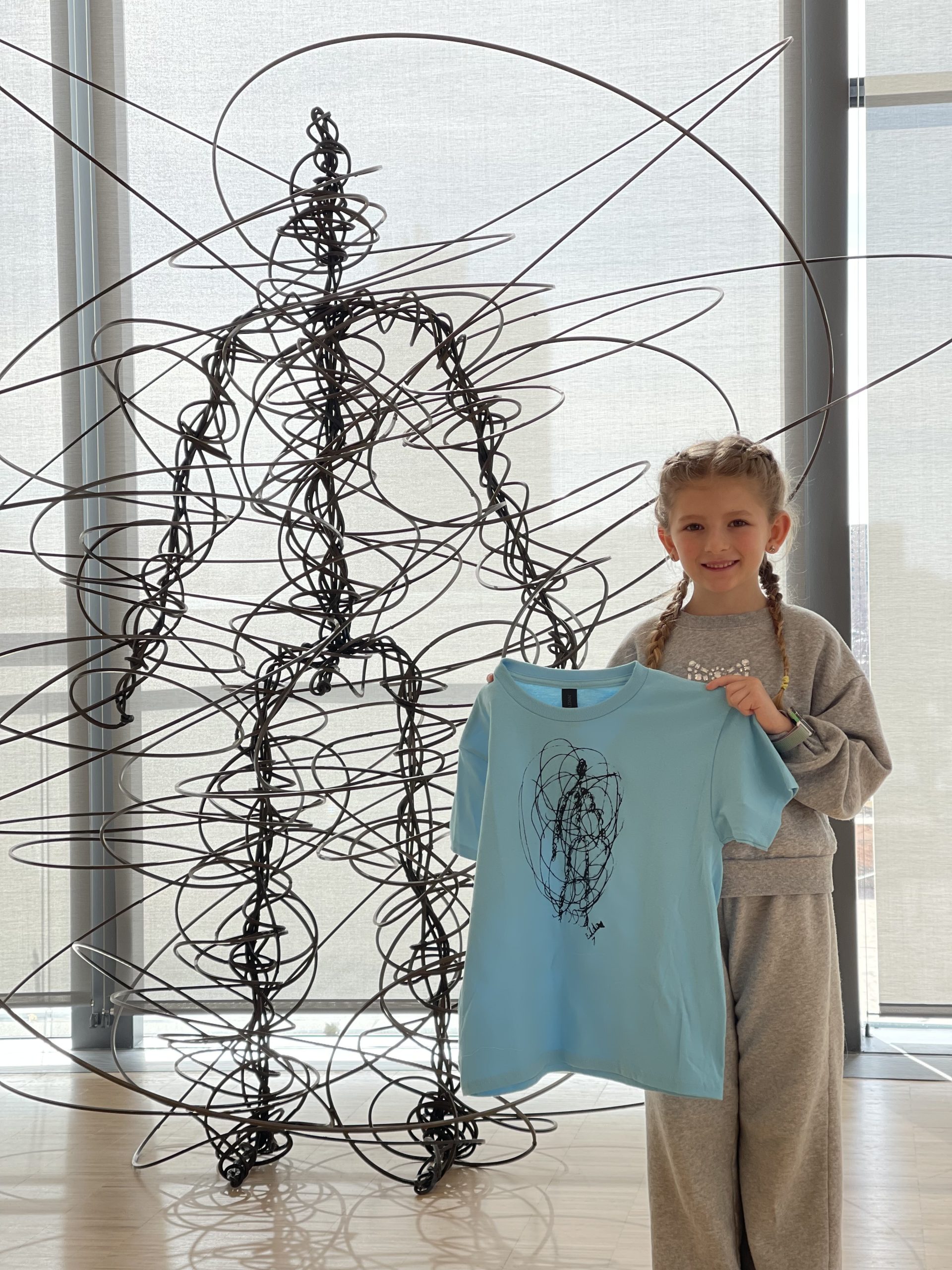 A smiling young girl with braided hair stands indoors, holding a light blue T-shirt featuring a wire figure design. Behind her is a large wire sculpture of a human figure, echoing the design on the shirt.