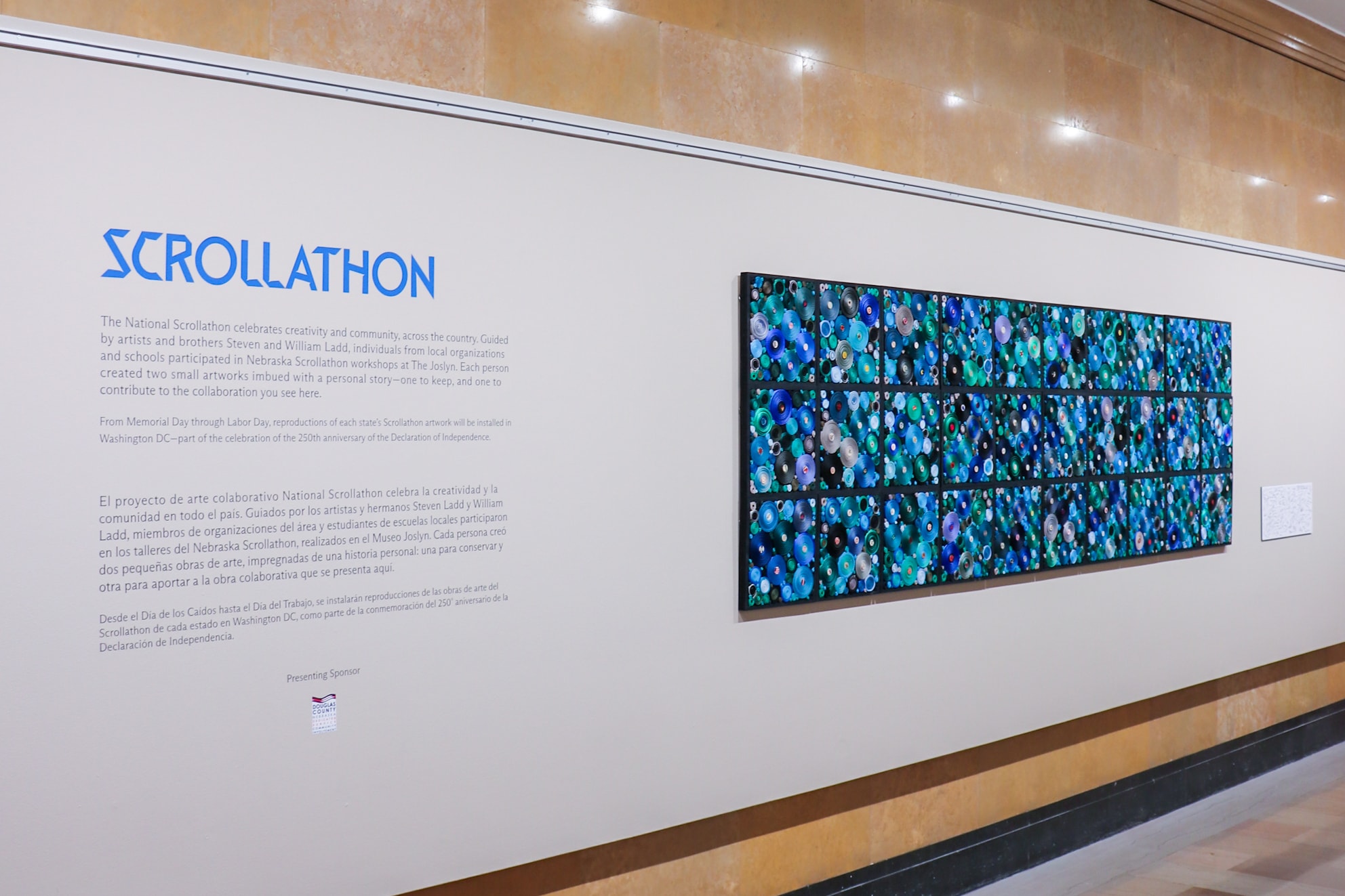 A museum wall displays a large text titled “Scrollathon” alongside a colorful, multi-panel artwork featuring abstract blue, green, and black circular patterns arranged in a rectangular grid.