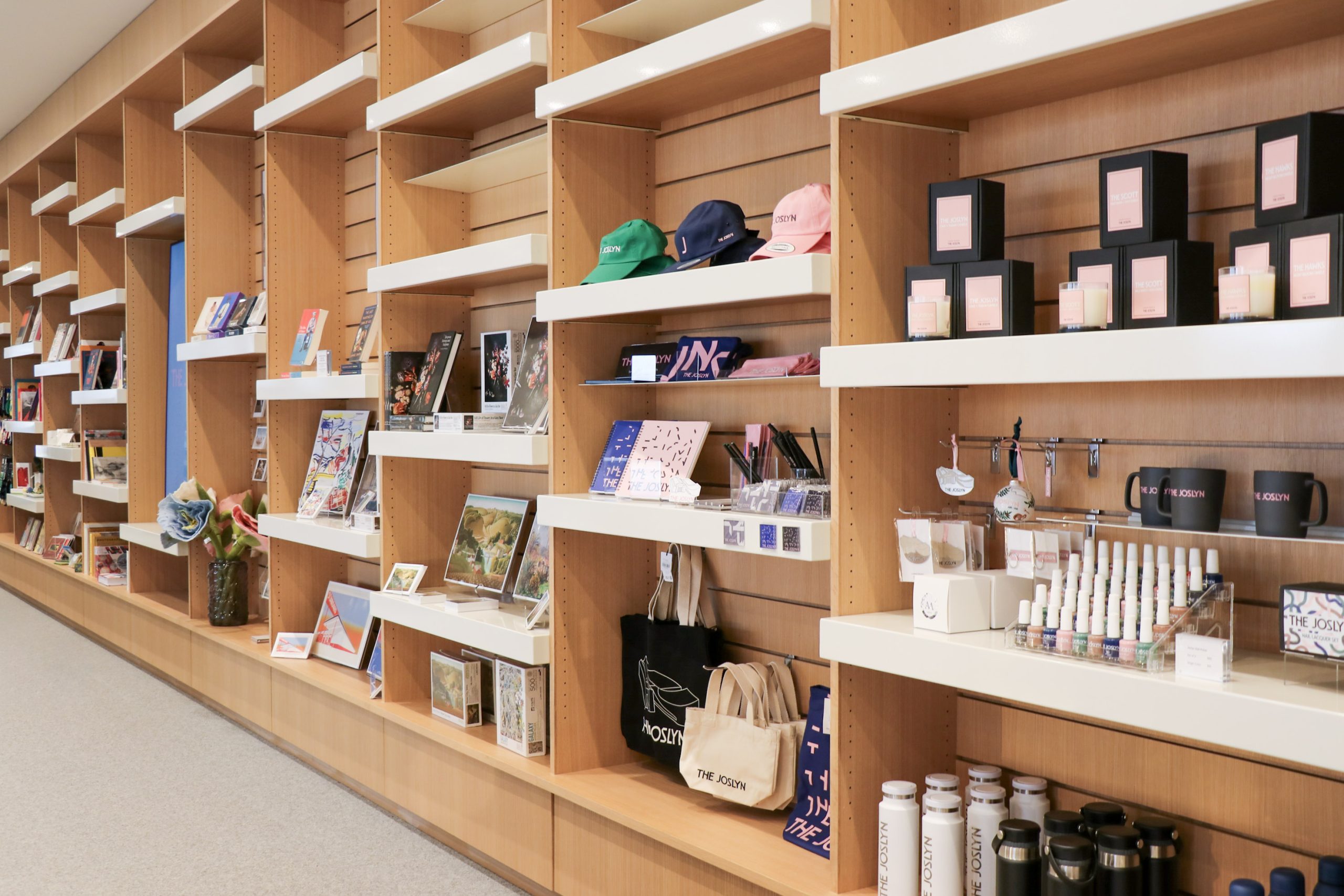 A modern gift shop display with wooden shelves holding various merchandise, including books, tote bags, caps, mugs, stationery, water bottles, framed art, and small gift items, all neatly arranged.