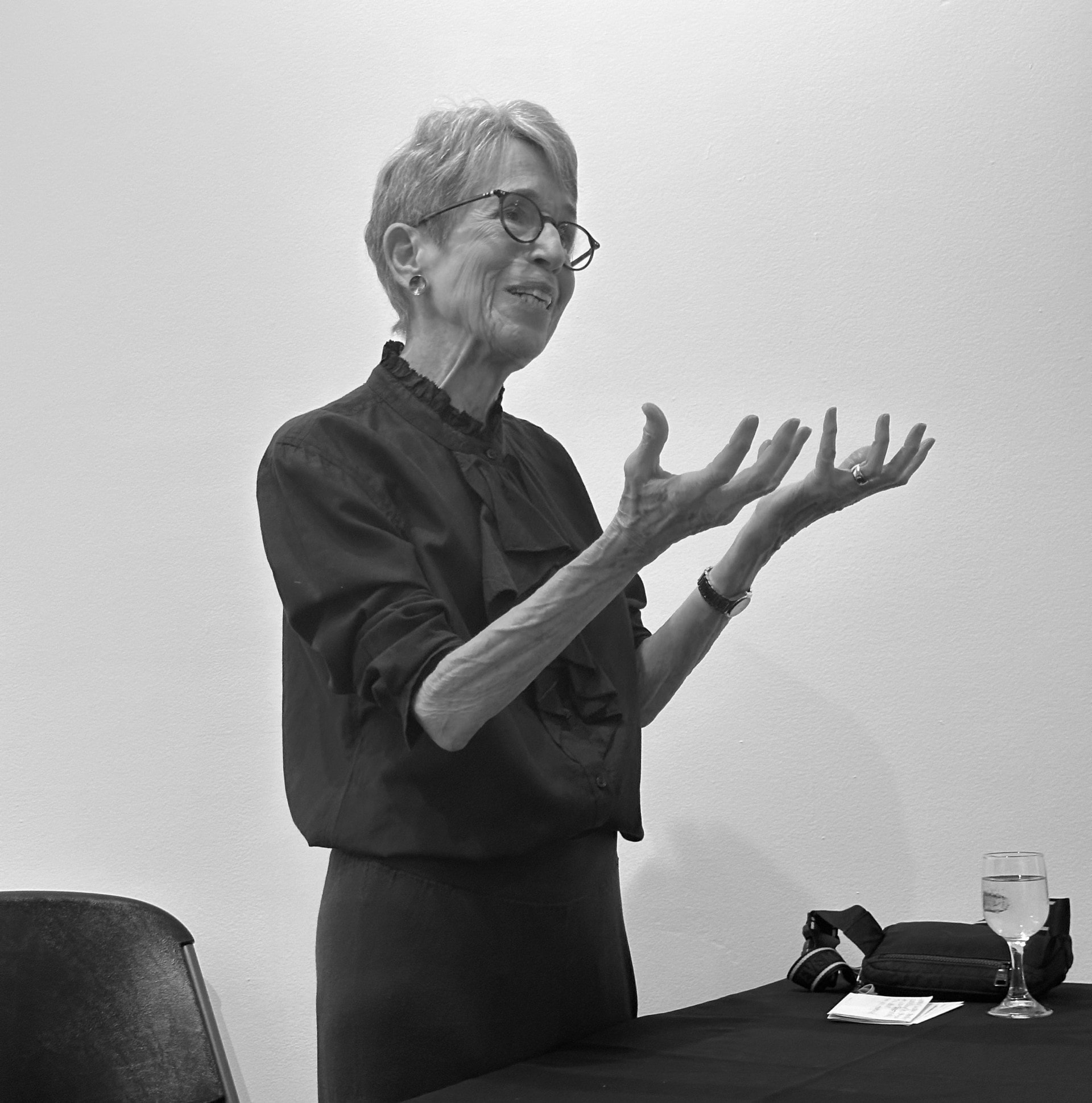 An older woman with short hair and glasses stands beside a table, gesturing expressively with both hands. She wears a dark blouse and appears to be speaking. A glass of water and a small bag are on the table in front of her.