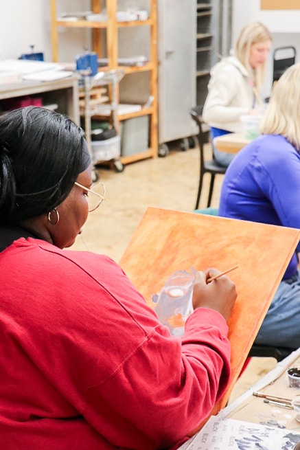 A woman in a red shirt paints on a canvas at a table in an art classroom. Other people are also seated and working on their own projects in the background.