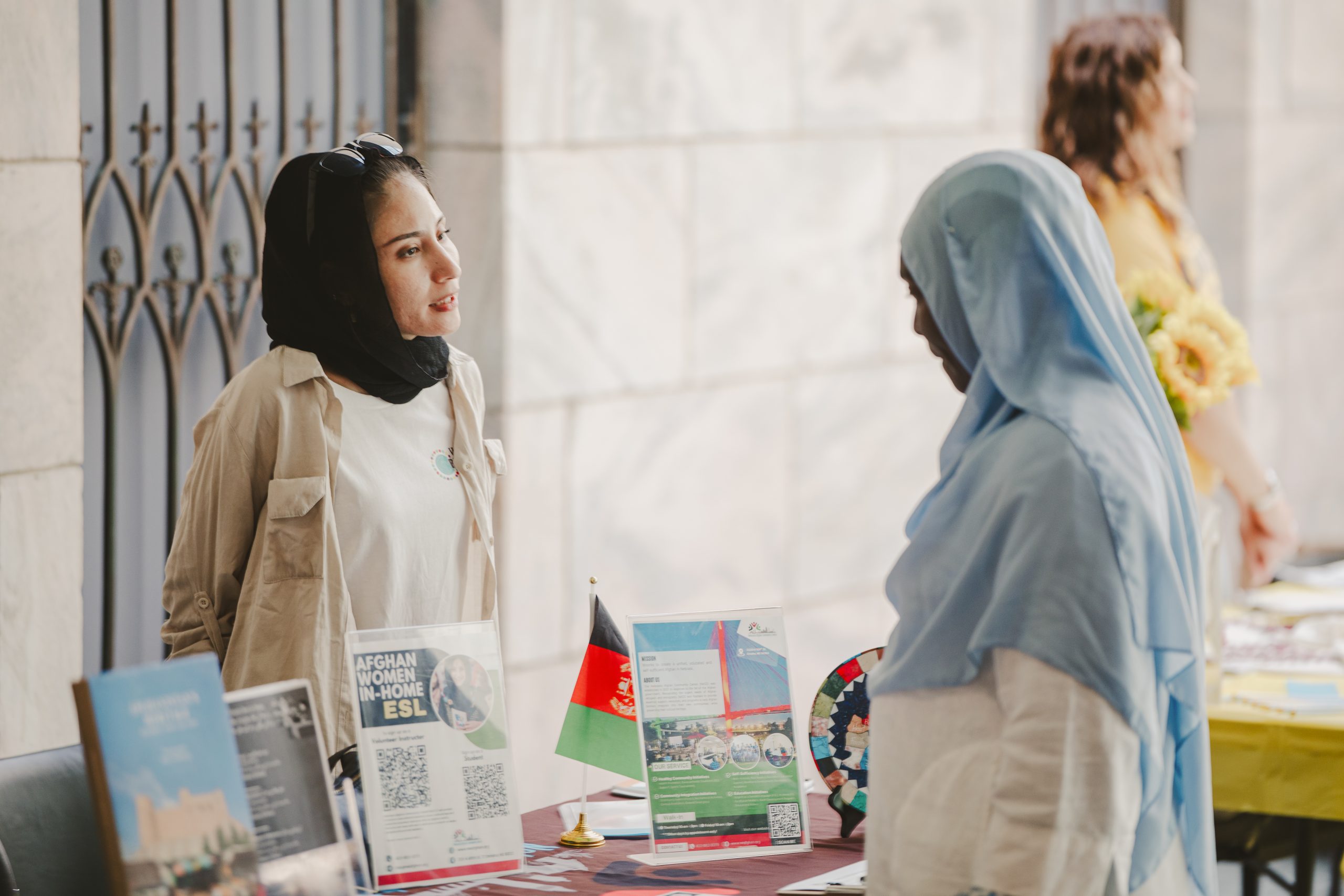 Two women wearing headscarves talk at a table with brochures, a small Afghan flag, and informational materials. Another person stands in the background holding a bouquet of yellow flowers.