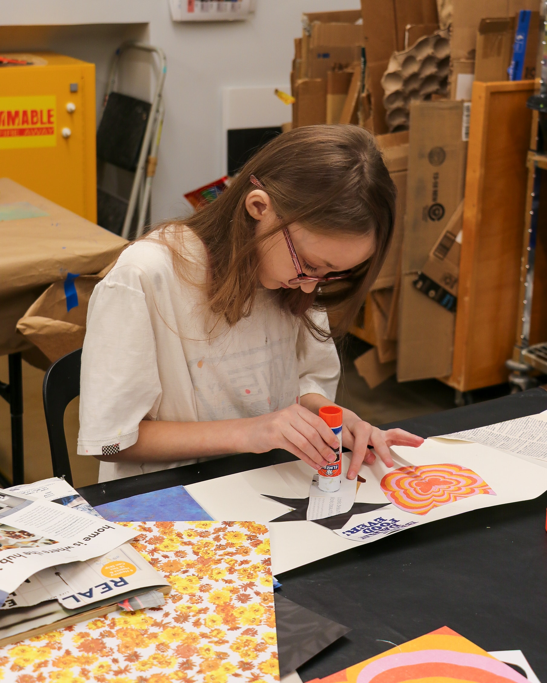A young girl with glasses sits at a table covered with colorful papers, using a glue stick to attach a piece of newspaper to her art project in a classroom or art studio setting.