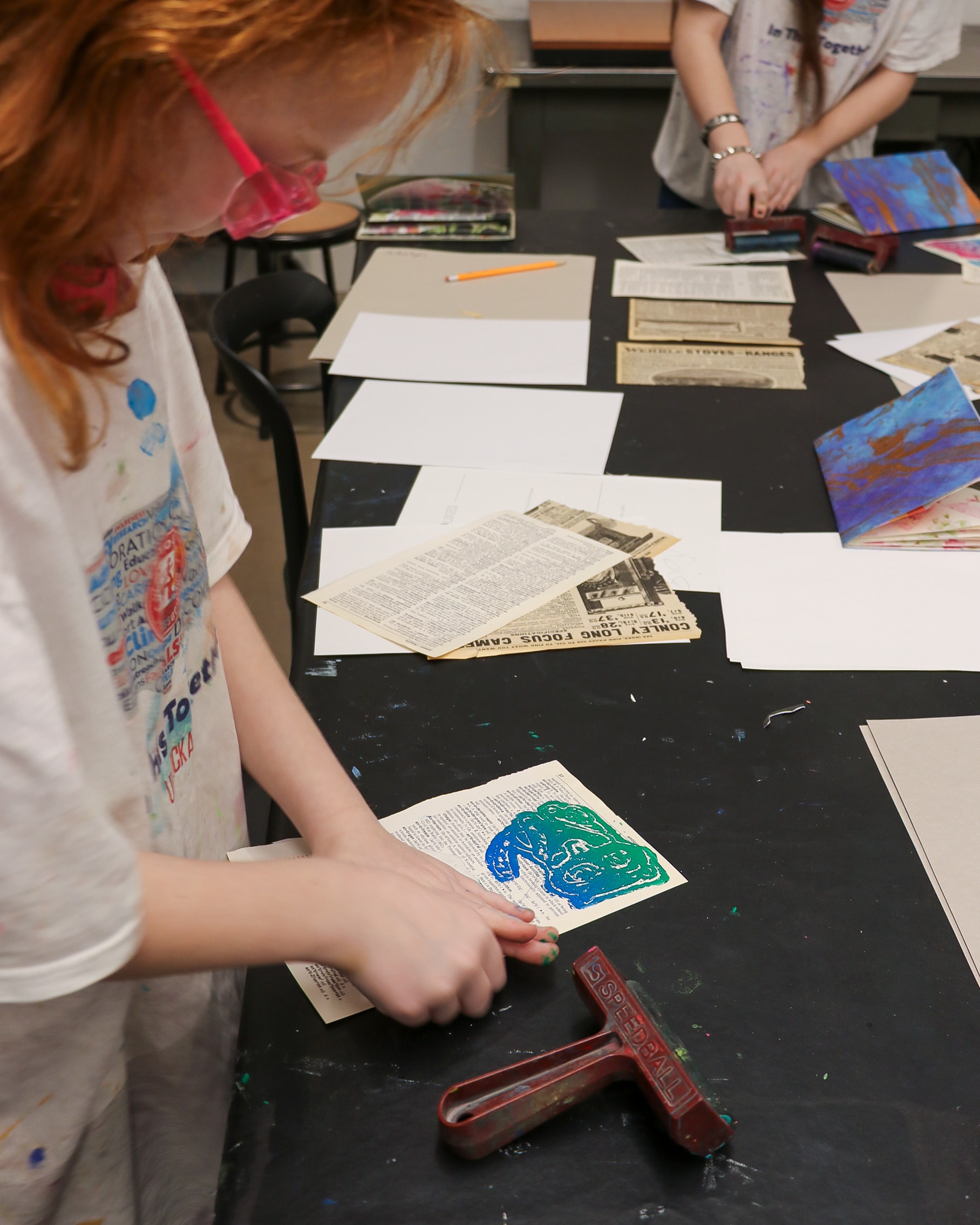 A child uses a red roller to apply blue and green paint to a book page on a black table. Art supplies, papers, and another person working are visible in the background.