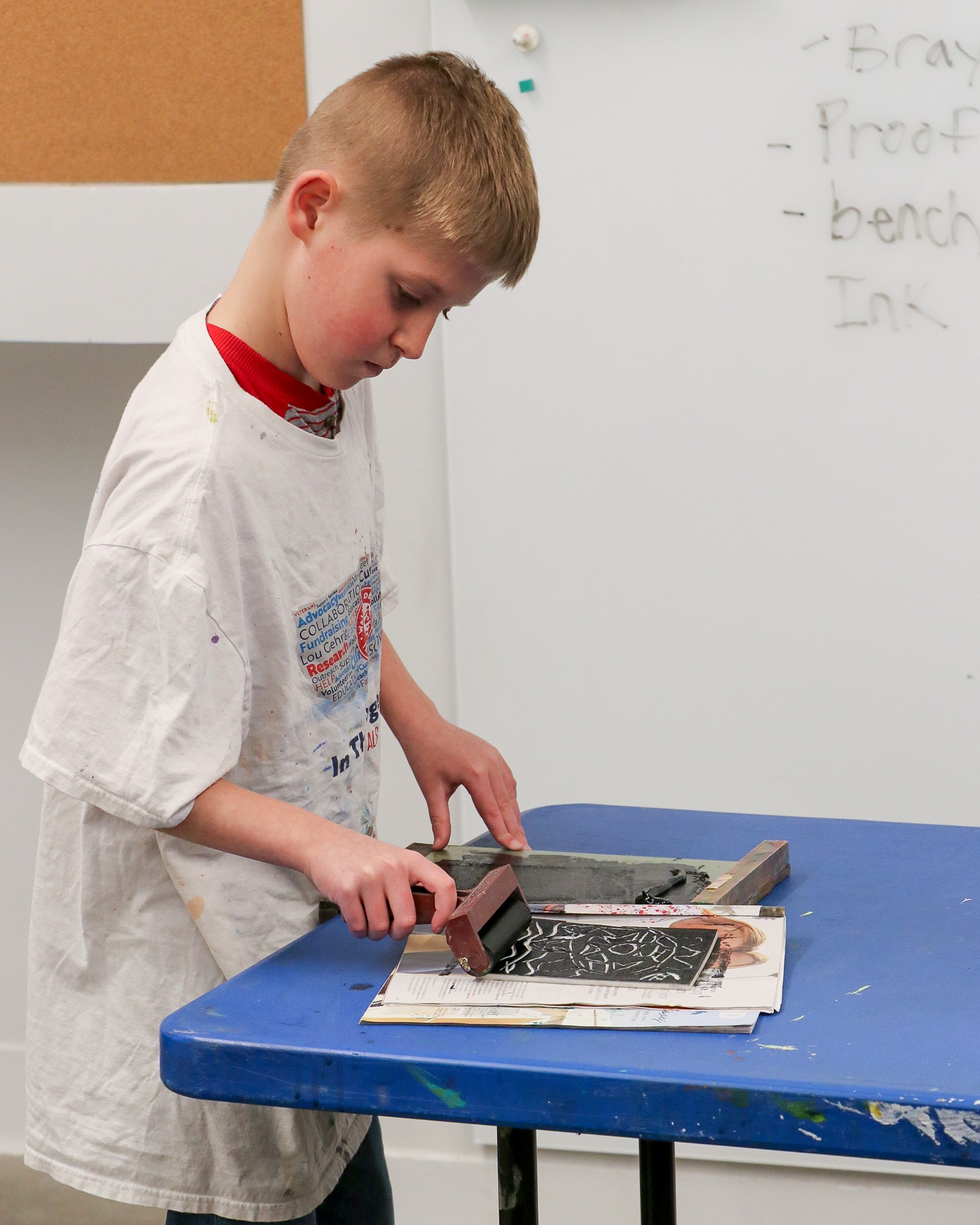 A boy in a paint-splattered shirt uses a brayer to roll ink onto a carved linoleum block, preparing for printmaking at a blue table with newspapers and tools.
