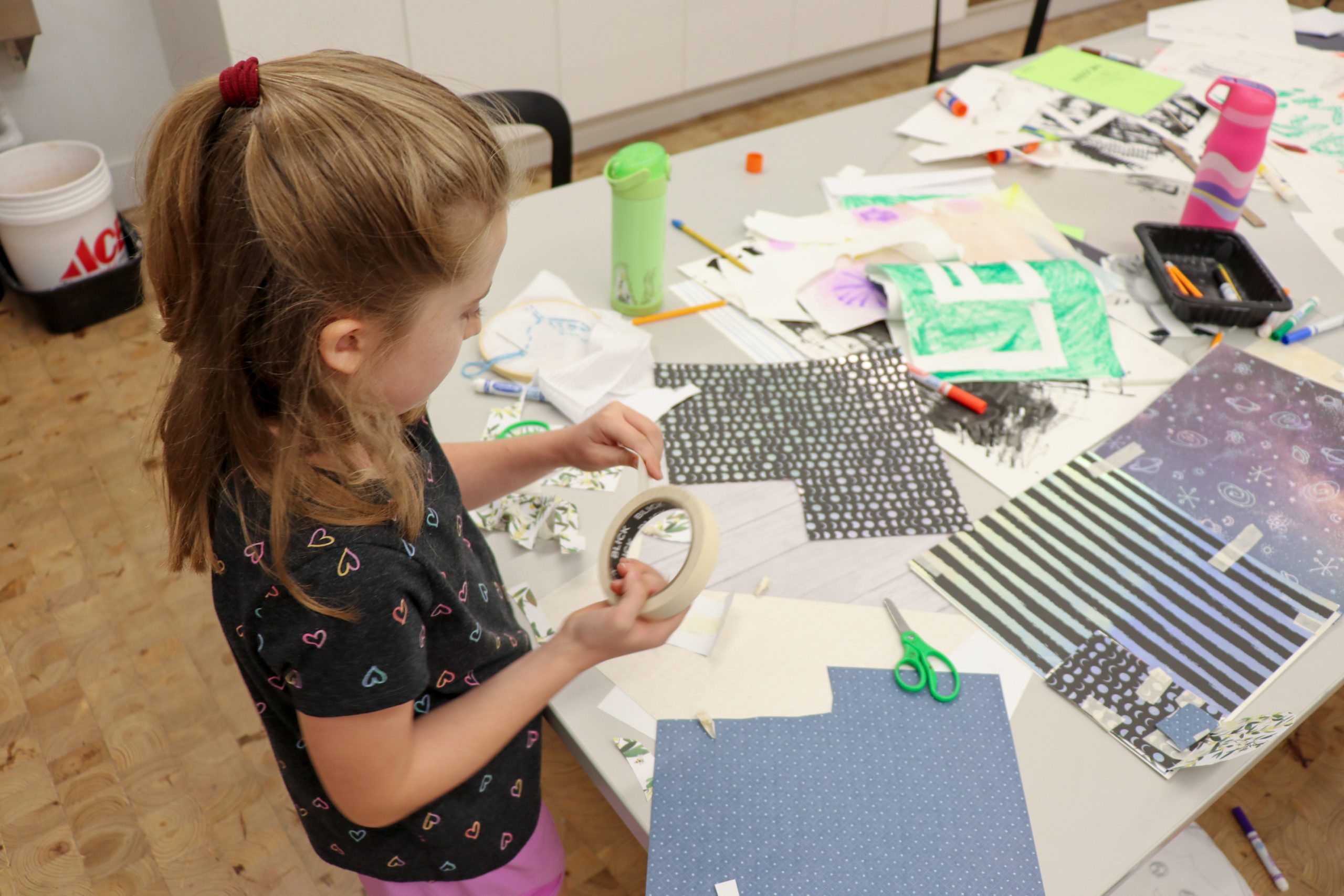 A young girl stands at a table covered with colorful papers, scissors, markers, and craft supplies. She holds a roll of masking tape, appearing focused on her creative project.