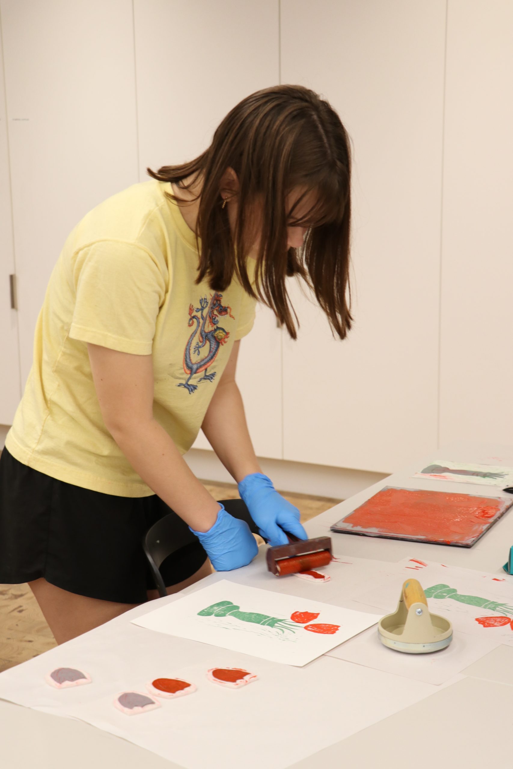 A girl wearing a yellow shirt and blue gloves uses a roller to apply paint to paper on a table, with art supplies and paint trays nearby in a brightly lit room.