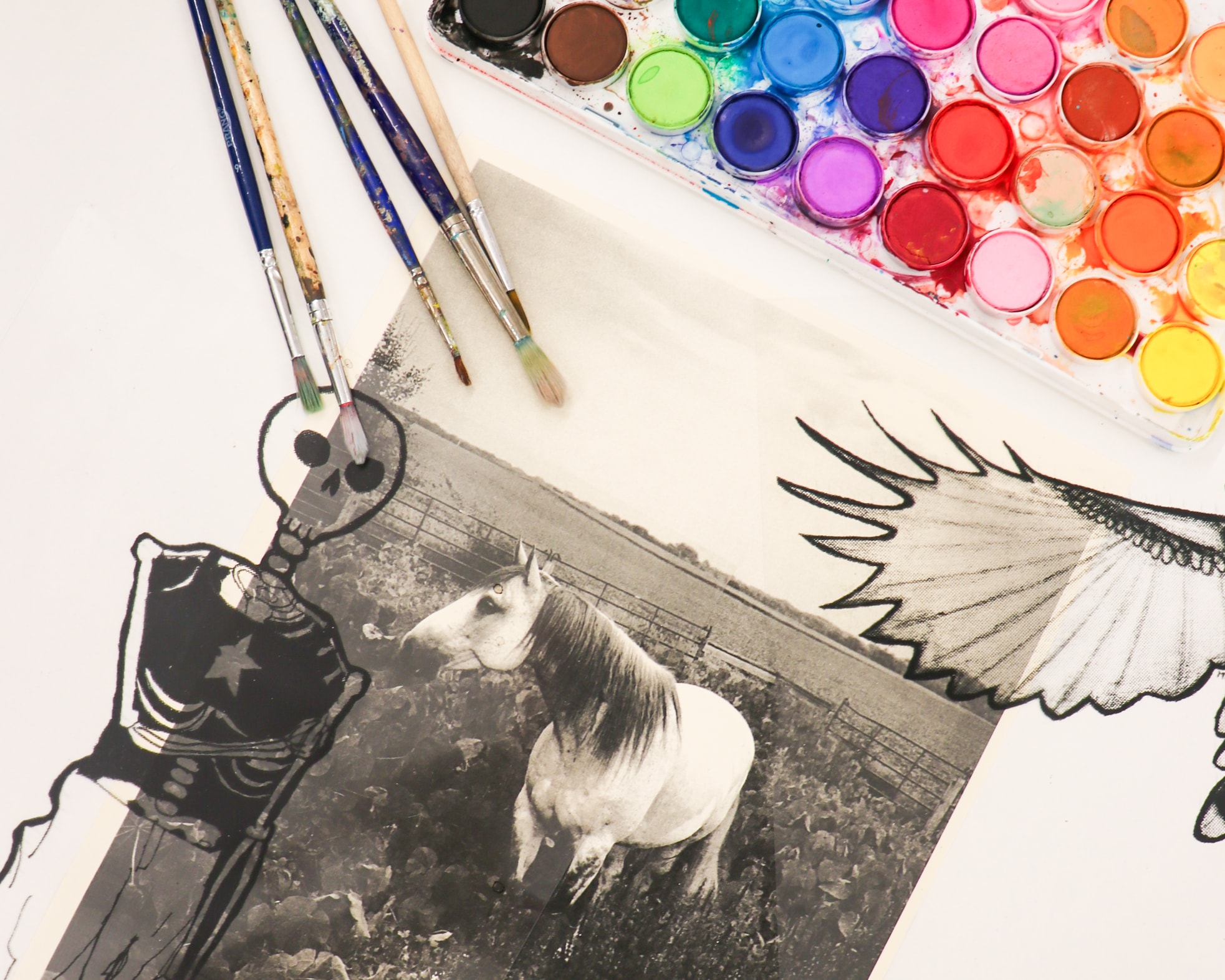 A black-and-white photo of a horse is surrounded by paintbrushes, a watercolor paint palette, and illustrated cutouts of a skeleton and a bird's wing on a white surface.