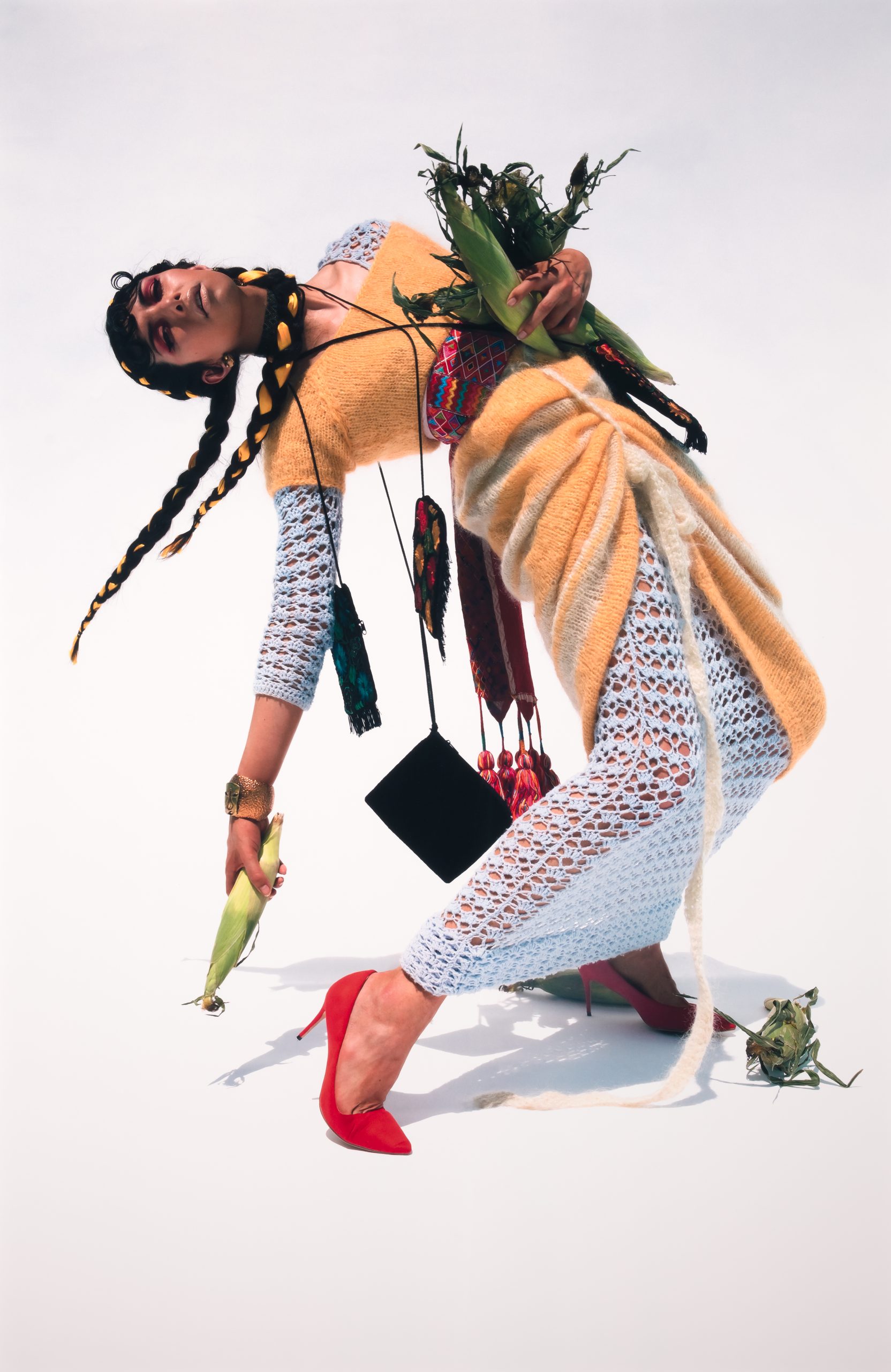 A person in a dramatic pose wears a pastel textured outfit, red heels, and braided hair with yellow ribbons. They hold corn cobs and accessories, with more corn and a frog at their feet, against a plain white background.