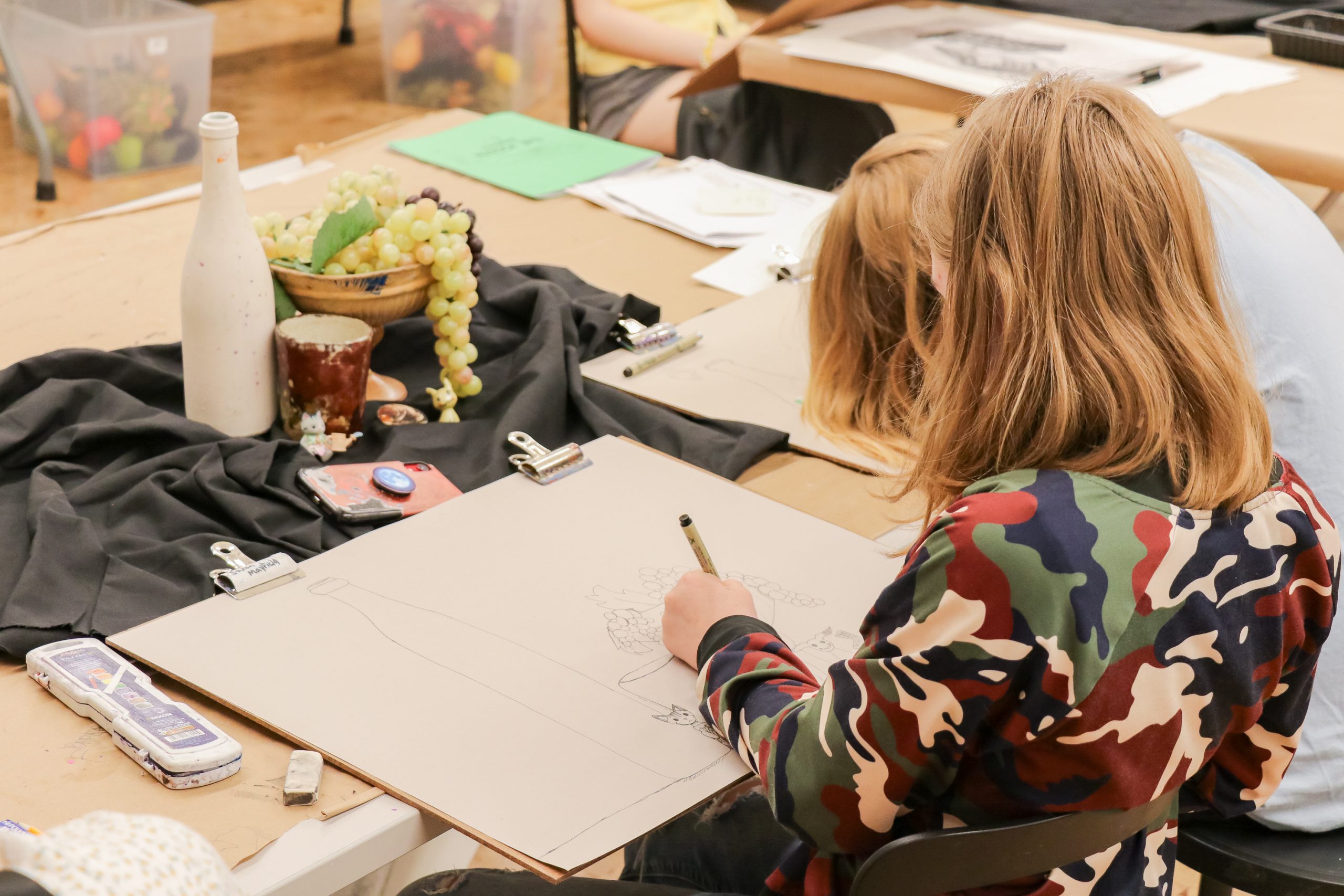 A person with long hair and a camouflage jacket sketches a still life of a bottle and fruit at a table, surrounded by art supplies and drawing materials. Other people are also working nearby.