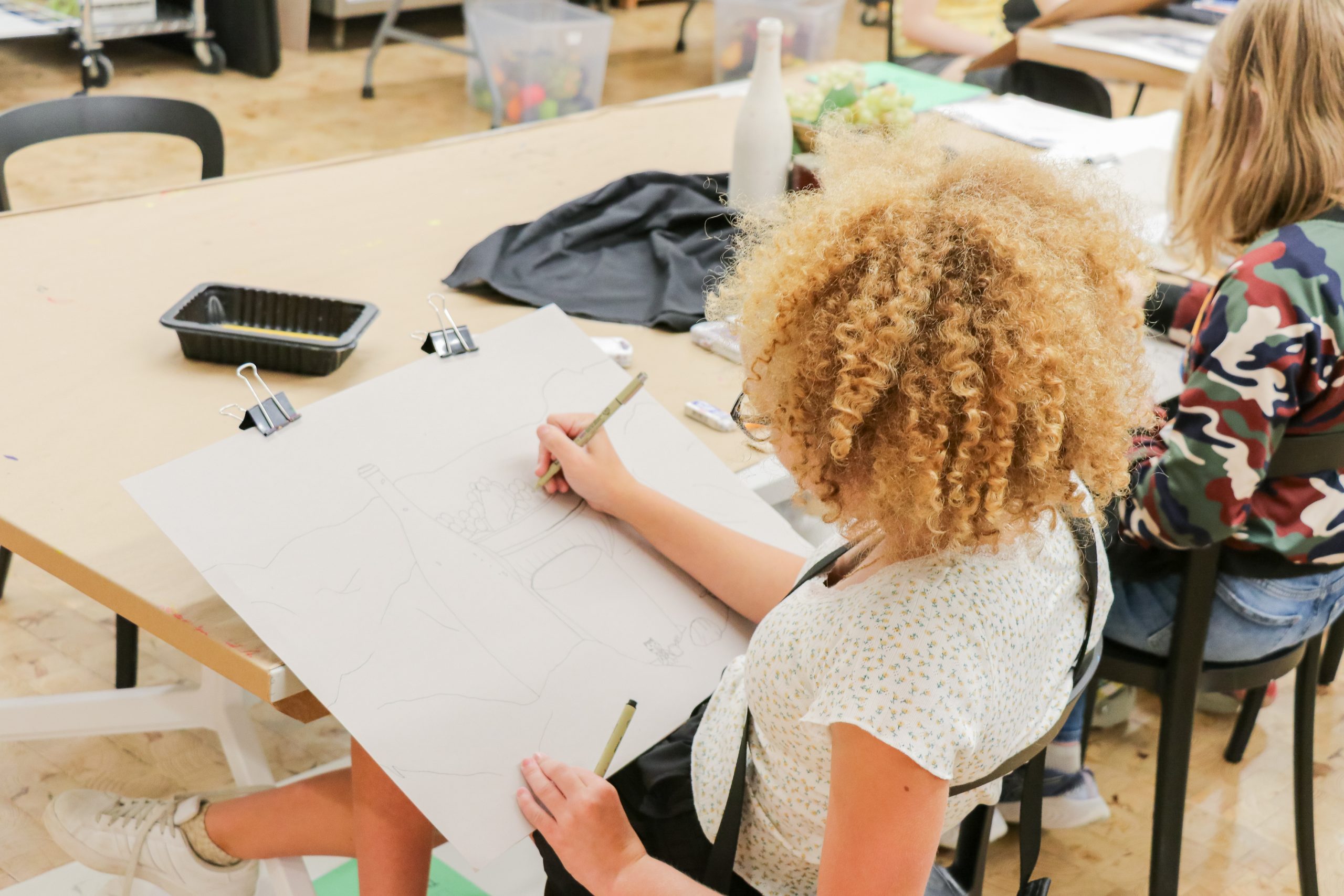 A child with curly hair sits at a table, sketching a still life drawing on a large sheet of paper in an art classroom. Art supplies and another student are visible in the background.