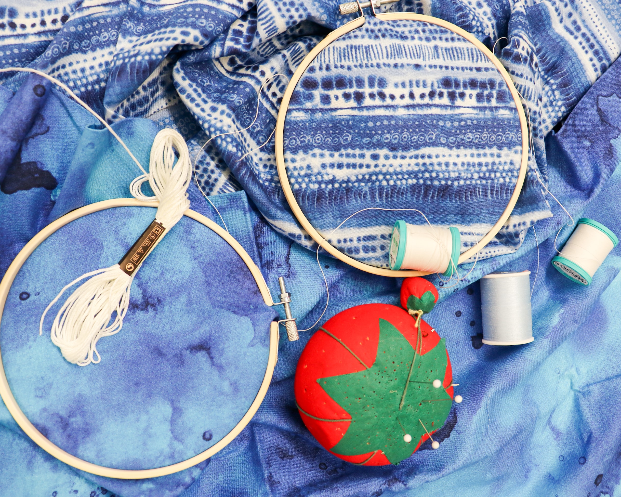 Two wooden embroidery hoops, white thread, a red pincushion with pins, and several spools of thread are arranged on patterned blue fabric.
