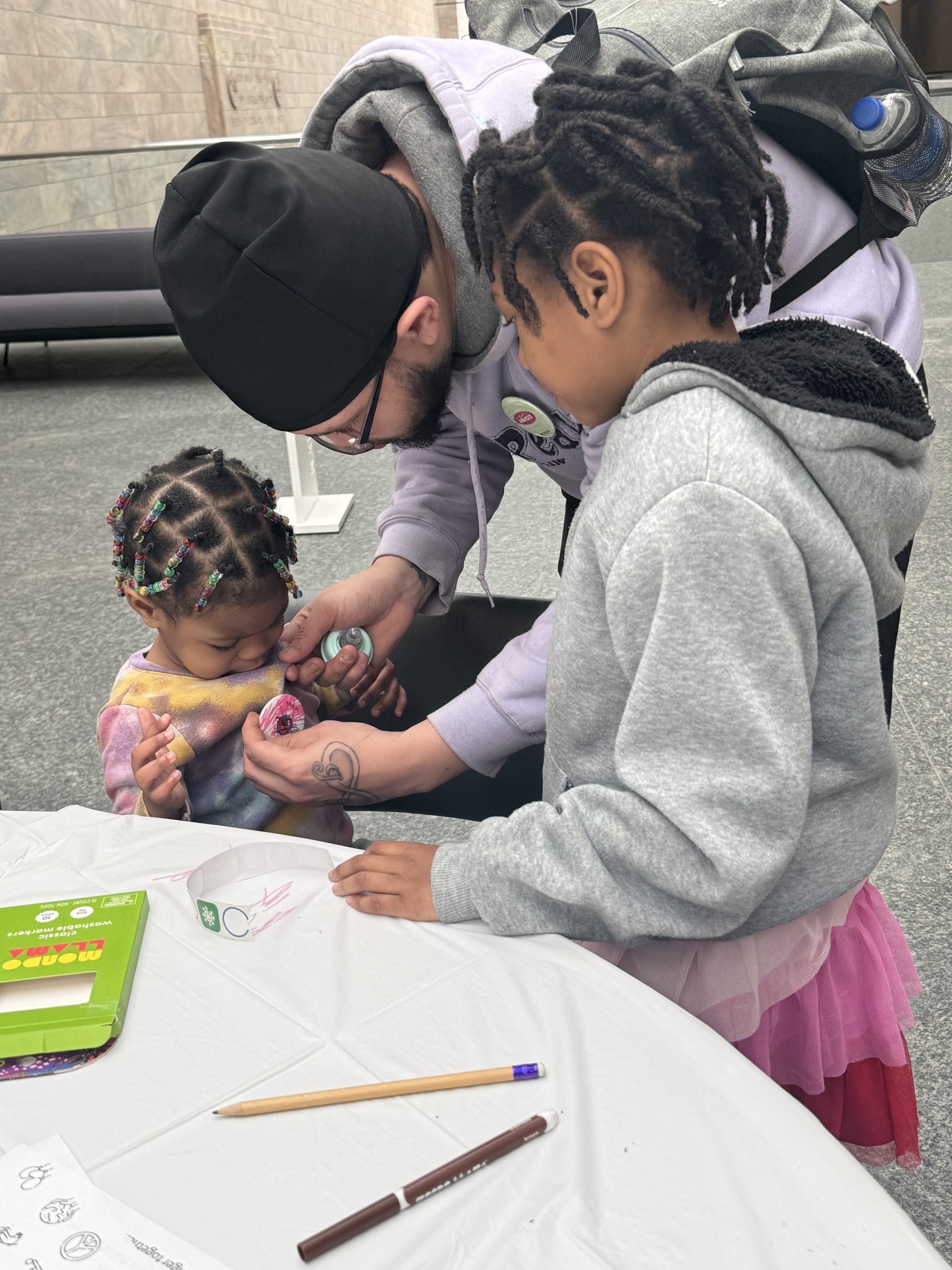 An adult helps a young child with beaded hair hold a marker while another child in a hoodie and tutu looks on. They are standing by a table with drawing supplies and papers.