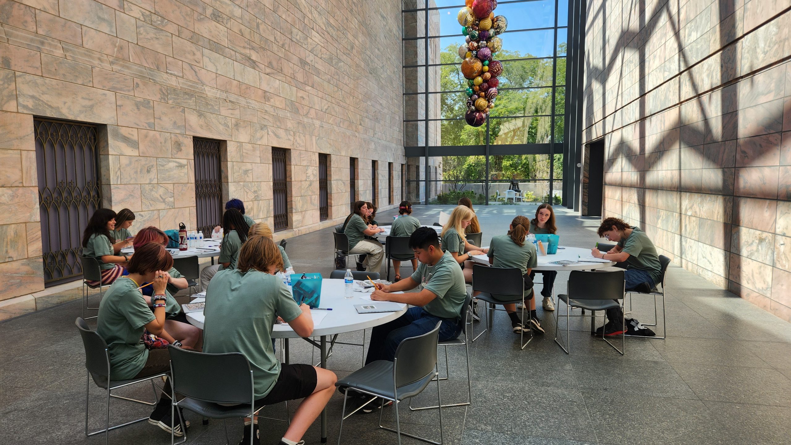 A group of people in matching green shirts sit at tables in a spacious, sunlit hall with stone walls, working on papers. Colorful glass art hangs near large windows at the end of the room.