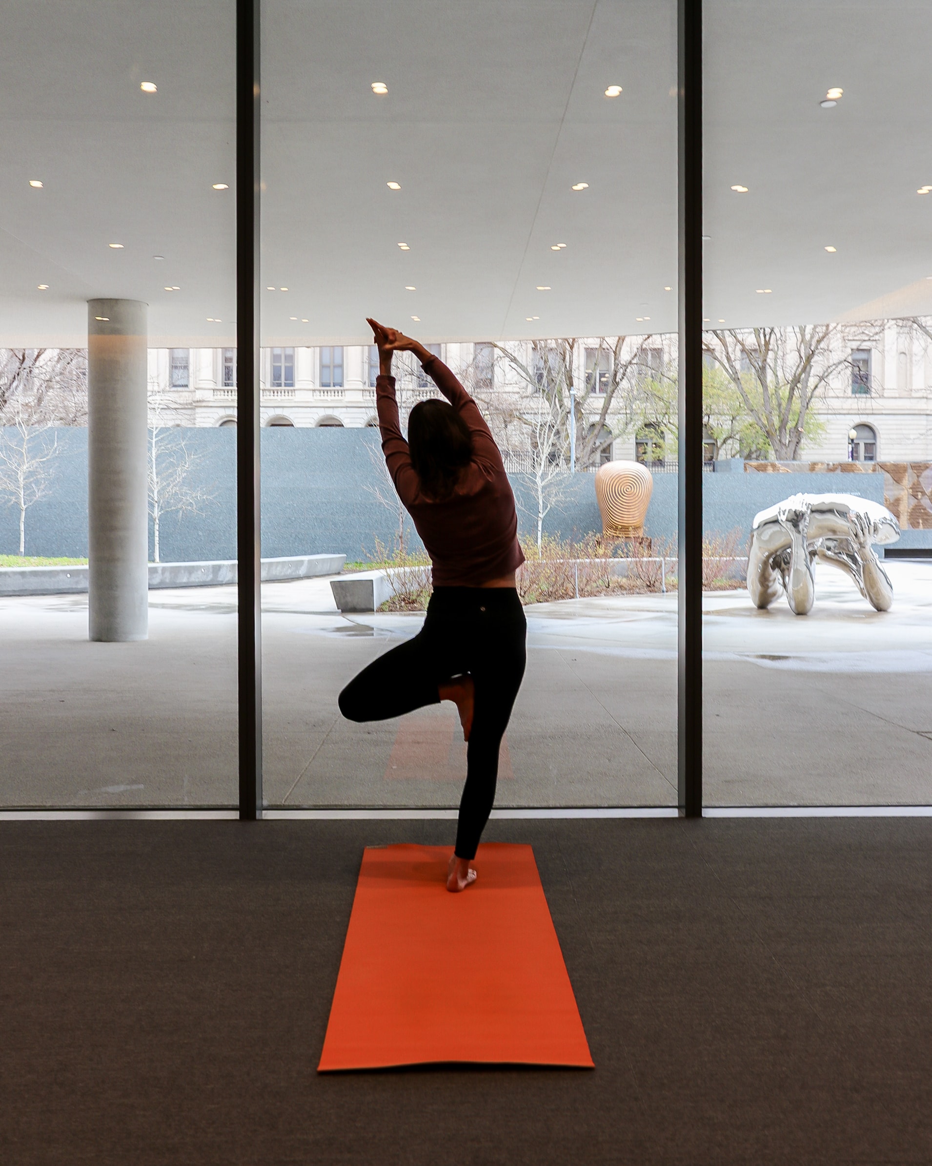 A person practices yoga on an orange mat indoors, standing in tree pose facing large floor-to-ceiling windows with a cityscape and outdoor sculptures visible outside.