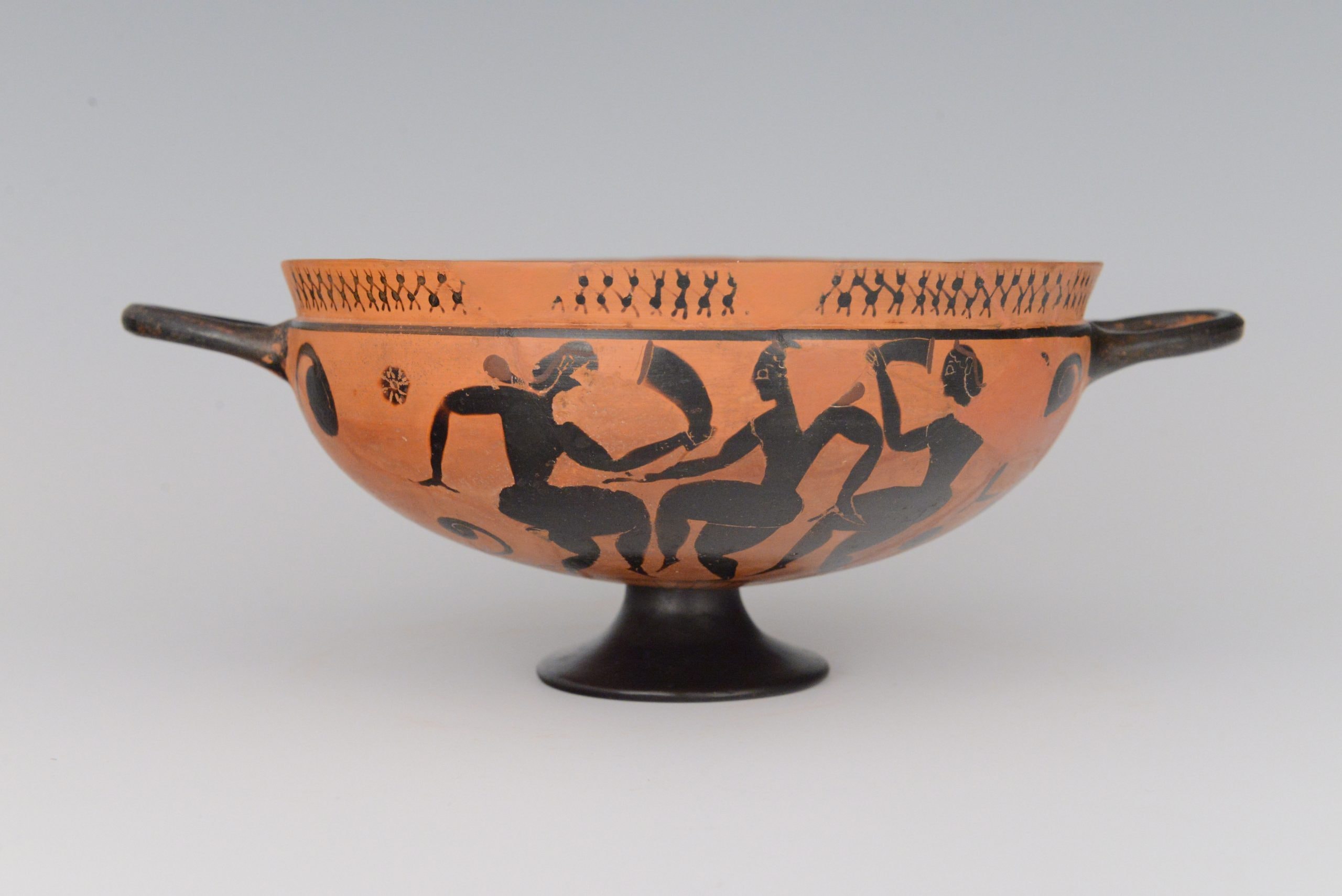 An ancient Greek terracotta drinking cup with two handles, decorated with black-figure artwork depicting human figures in dynamic poses, set against an orange background.